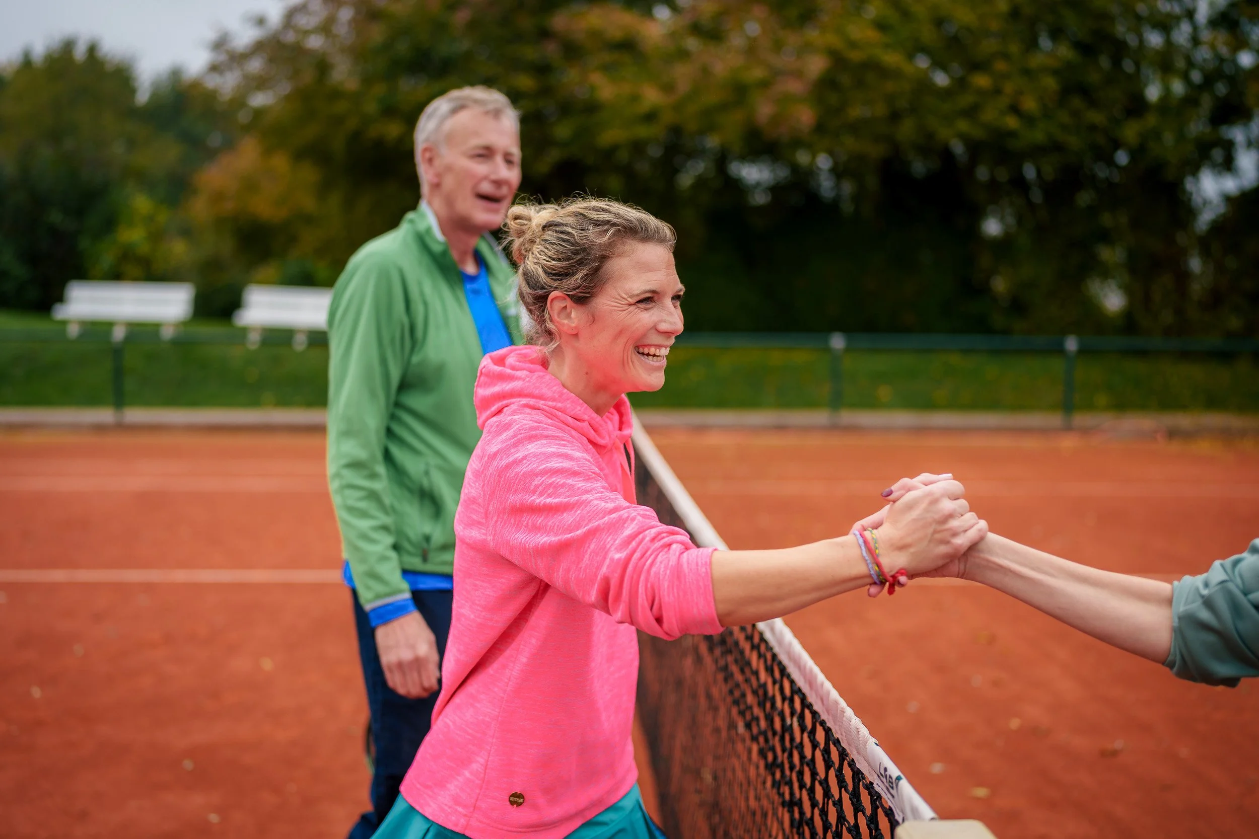 Zwei lachende Erwachsene am Tennisnetz, die sich die Hand geben, während im Hintergrund ein weiterer Mann auf dem Tennisplatz steht, mit Bäumen im Herbst im Hintergrund.