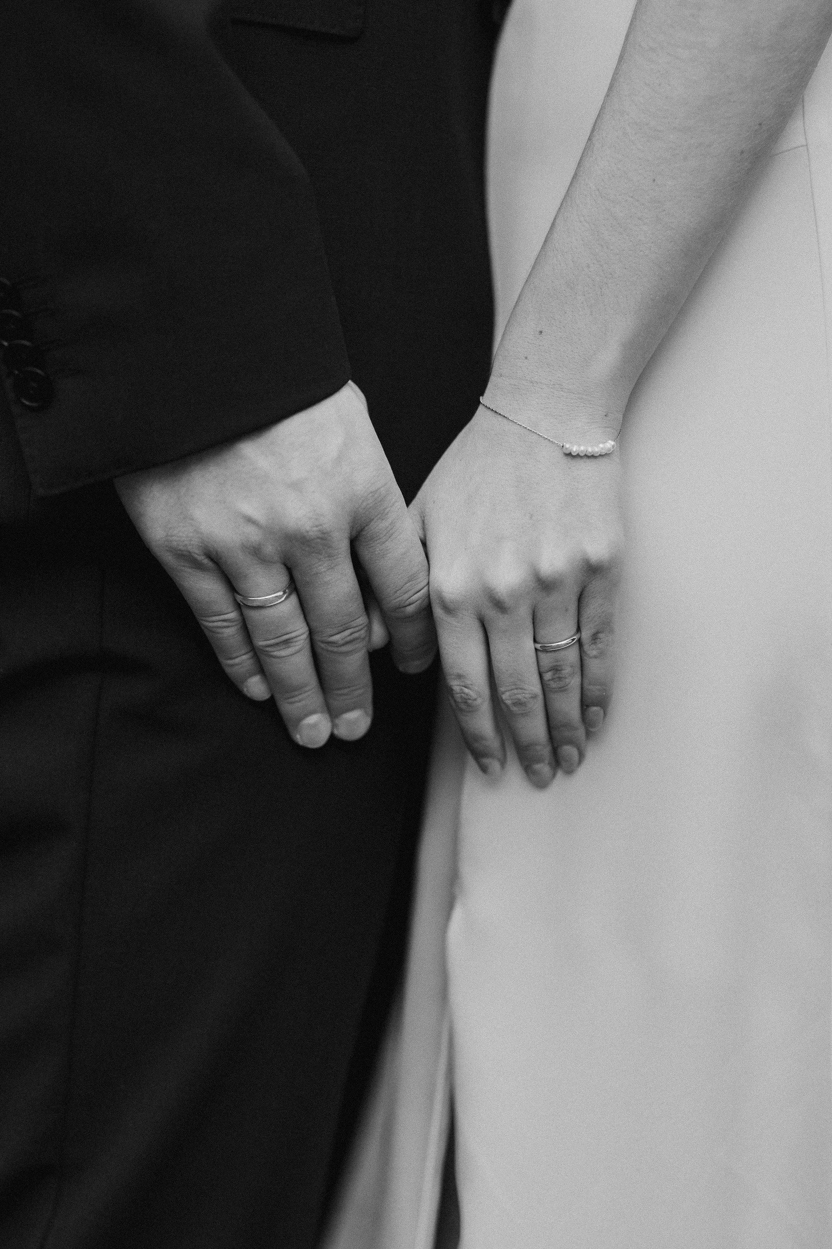 Nahaufnahme von den Händen eines Paares, die bei einer Hochzeitszeremonie gehalten werden, beide tragen Eheringe, die Frau trägt eine Armbanduhr und ein dünnes Armband.