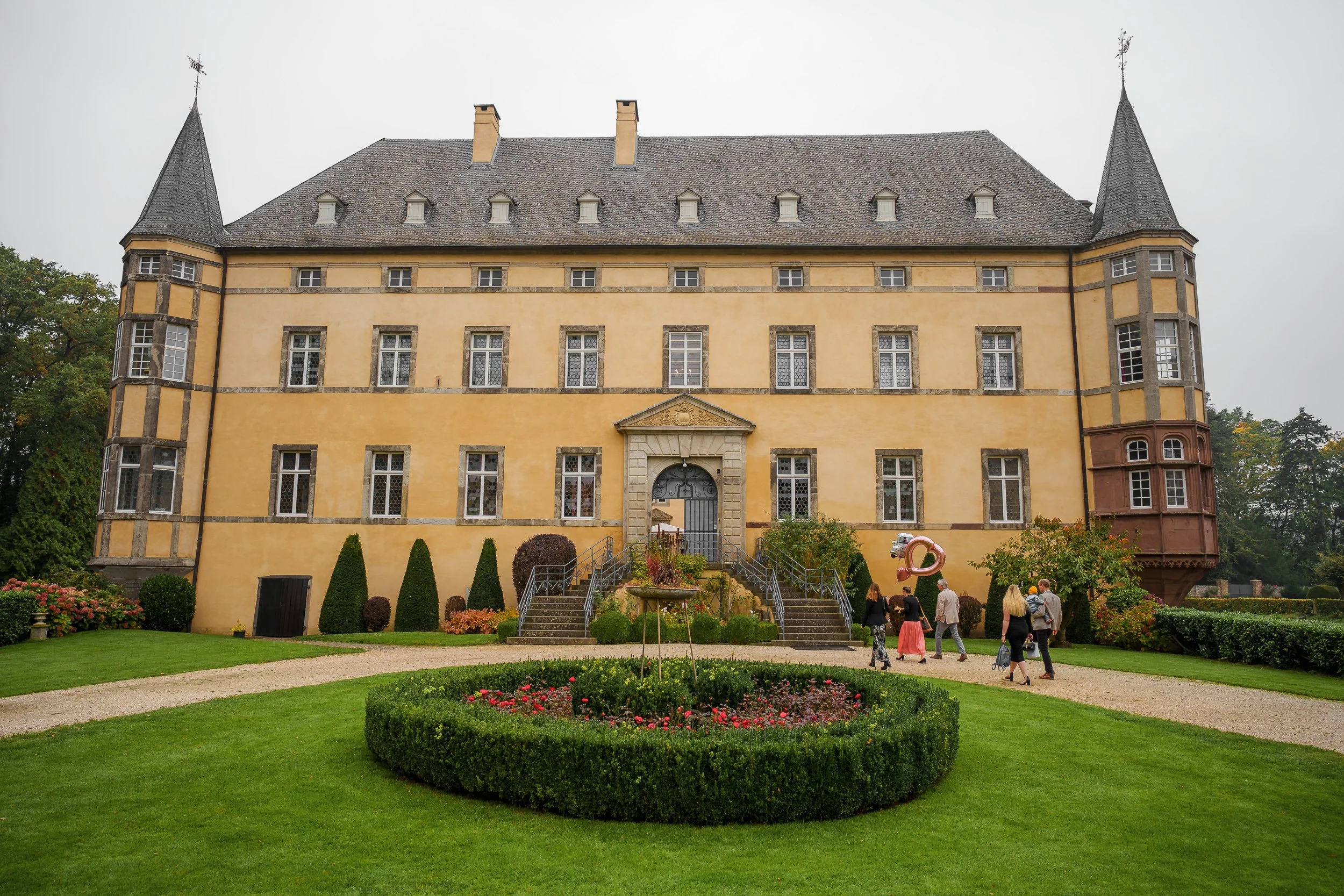 Ein historisches Schloss mit gelber Fassade, mehreren Fenstern und spitzen Türmen, umgeben von einem gepflegten Garten mit Blumen, Gehwegen und Büschen, an einem bewölkten Tag.