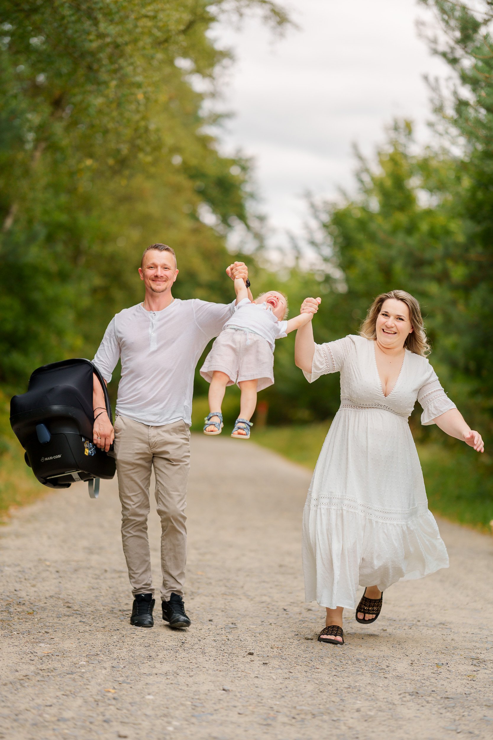 Familie mit zwei Erwachsenen und einem Kind, die auf einem Weg mit Bäumen im Hintergrund spazieren. Das Kind wird in die Luft gehalten, alle lachen und scheinen Spaß zu haben.