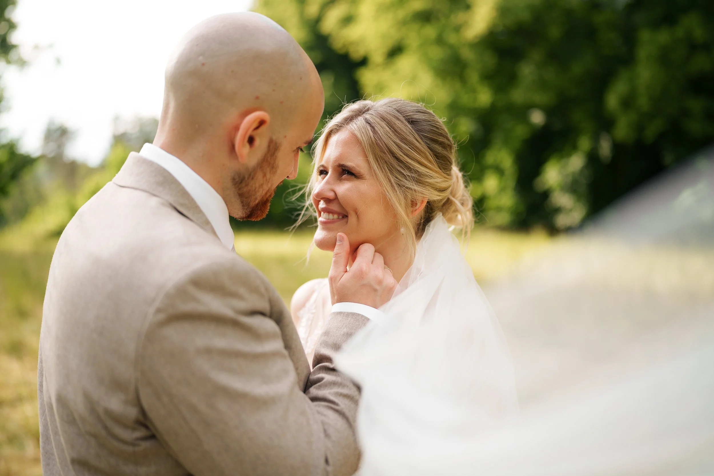 Ein Mann und eine Frau bei einer Hochzeit im Freien, der Mann berührt das Kinn der lachenden Frau, im Hintergrund Bäume und eine Wiese.