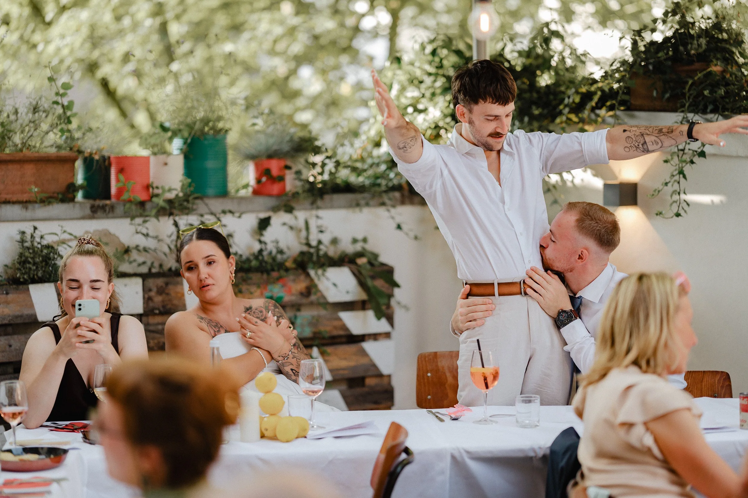 Menschen feiern an einem Tisch, bei einer Veranstaltung im Freien. Zwei Männer stehen in der Mitte, einer küsst den anderen auf die Brust, während der andere die Arme weit ausstreckt. Drei Frauen sitzen um den Tisch herum, zwei schauen in Richtung de