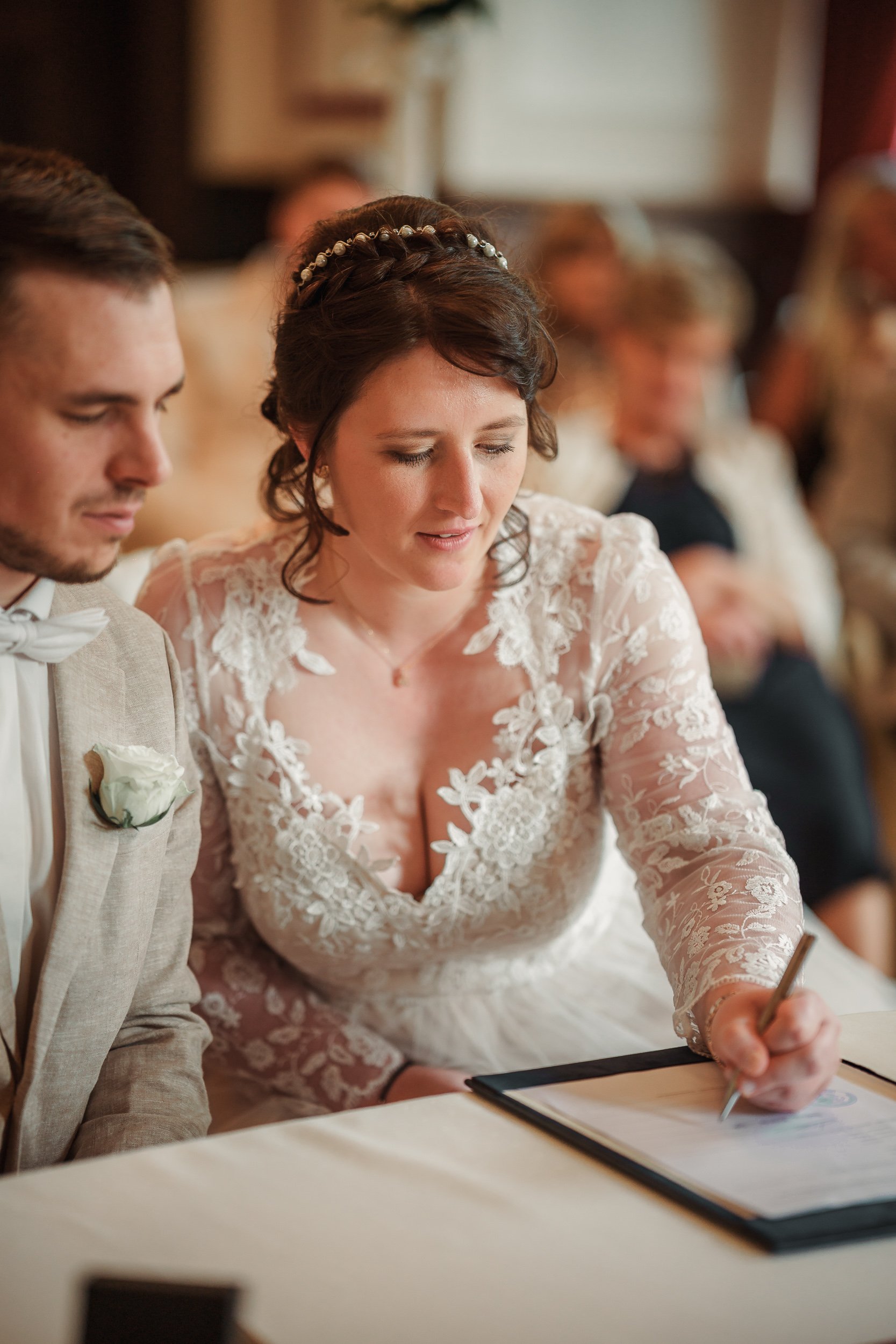 Braut und Bräutigam bei der Unterschrift der Hochzeitsdokumente, die Braut trägt ein Spitzenkleid mit tiefem Dekolleté und der Bräutigam einen hellen Anzug mit Krawatte.