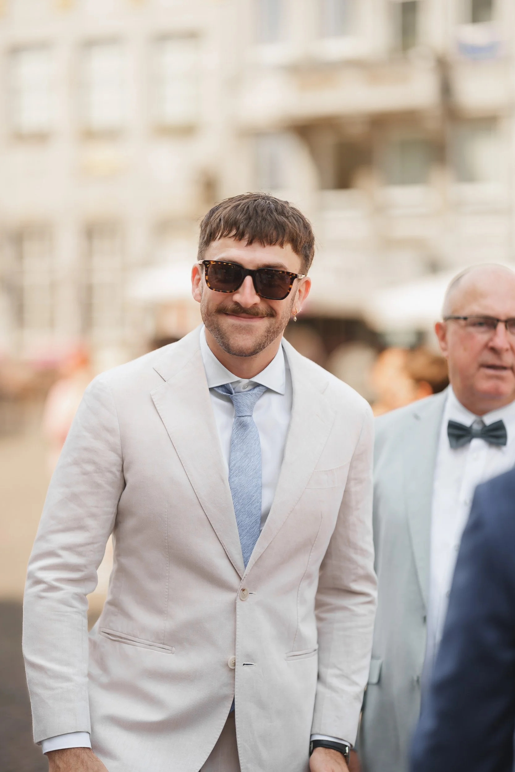 Mann in weißem Anzug, blauer Krawatte und Sonnenbrille bei einer formellen Veranstaltung im Freien.