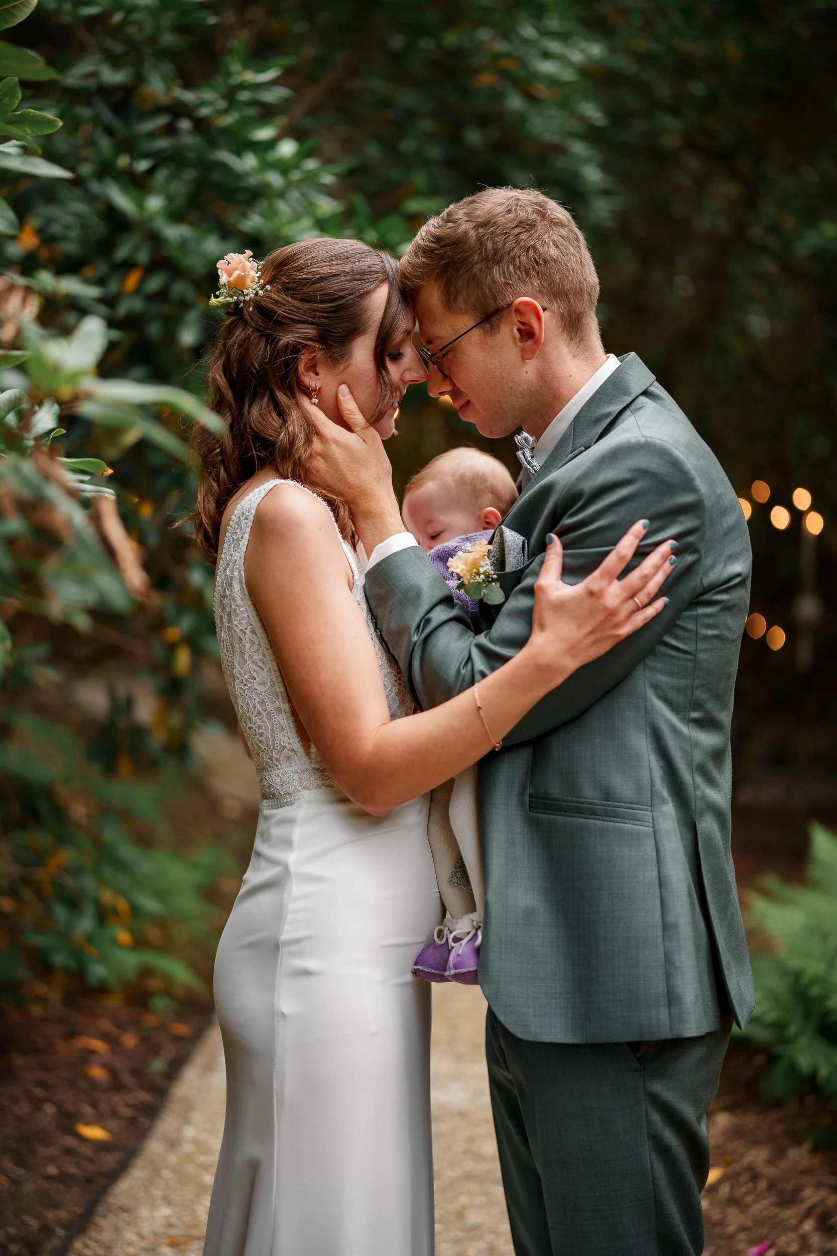 Ein heiratendes Paar in einem Garten, sie berühren sich mit ihren Stirnen, während der Mann ein Baby hält, das auf ihrer Brust schläft.