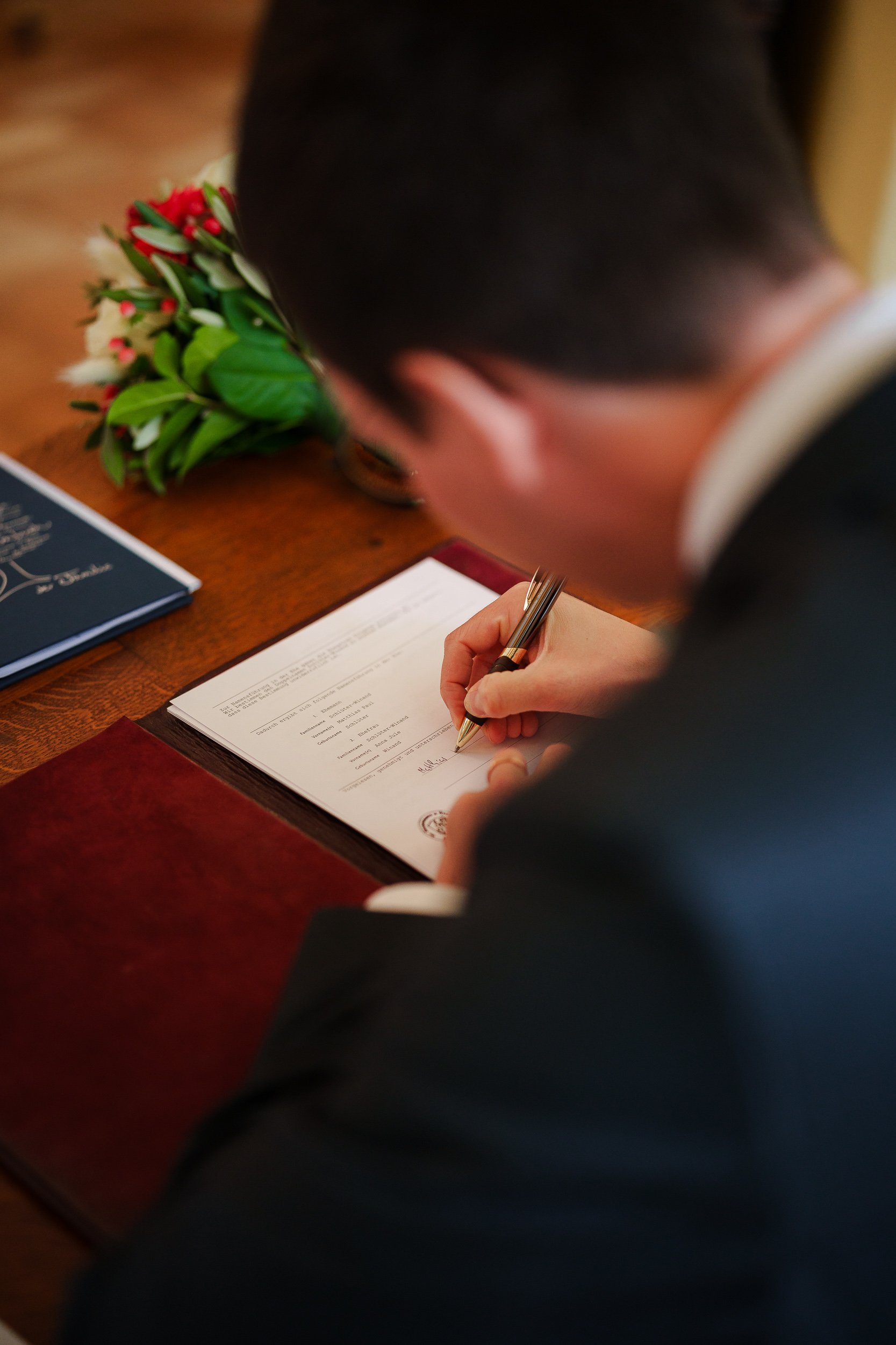 Ein Mann unterschreibt ein Dokument auf einem Schreibtisch. Neben ihm liegt ein Blumenstrauß und ein Buch.