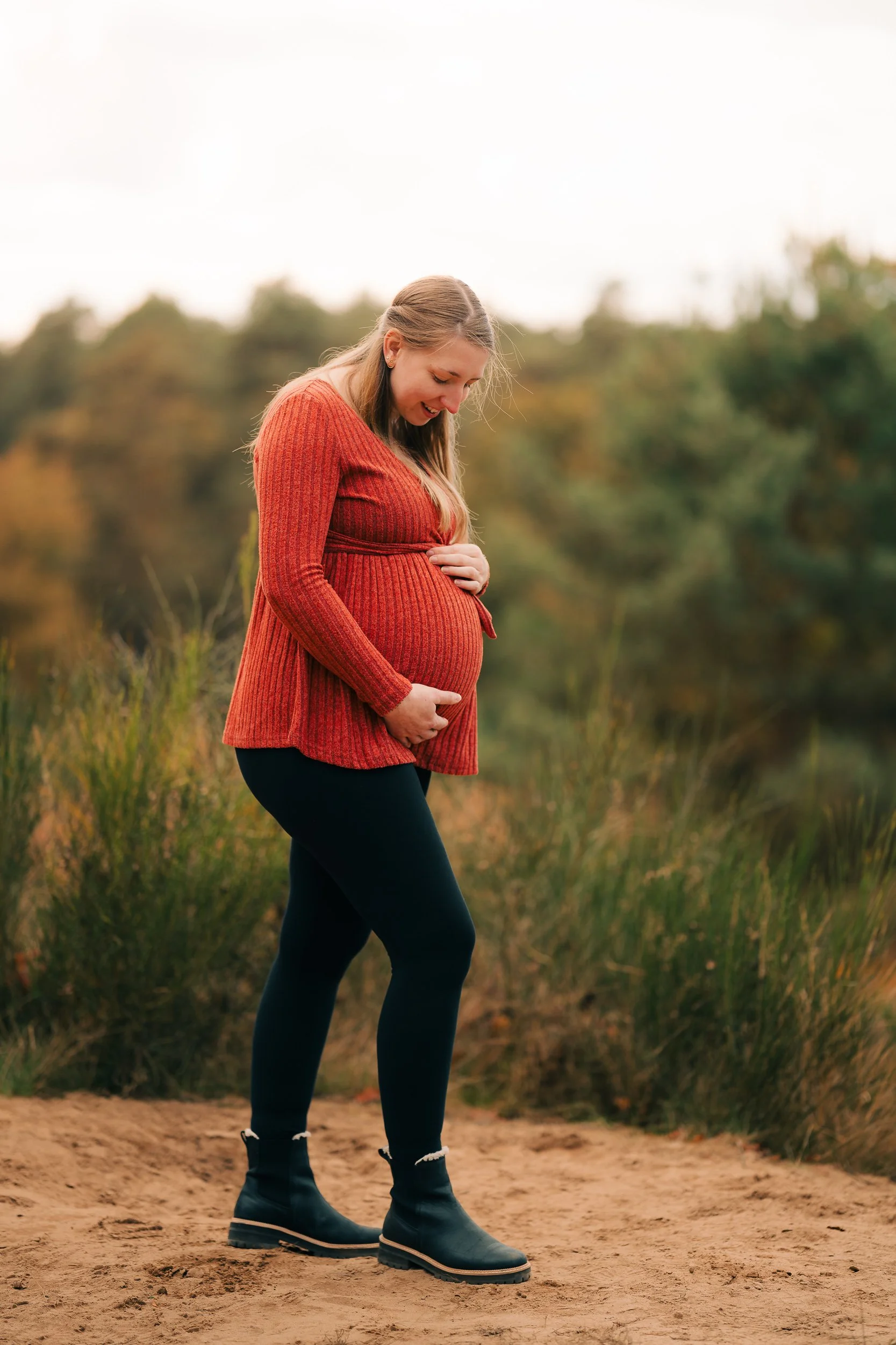 Schwangere Frau in einem roten Pullover und schwarzen Leggings hält ihren Babybauch und schaut lächelnd nach unten, während sie auf einem Naturpfad steht.