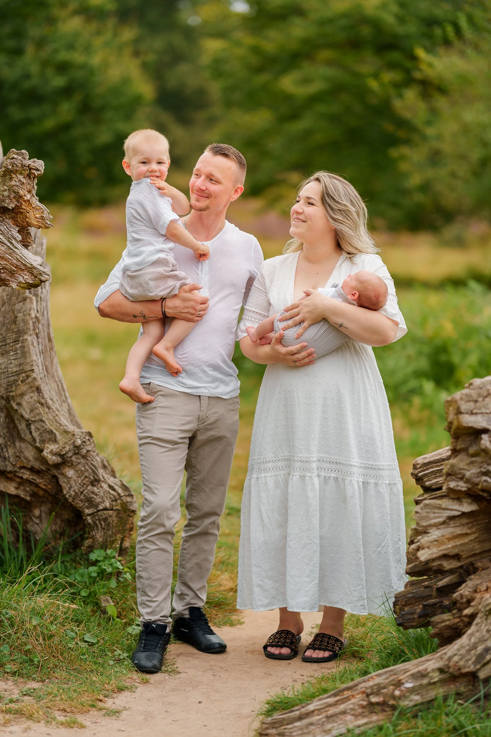 Familie mit Mutter, Vater, einem Kleinkind und einem Baby im Freien auf einem Waldweg, umgeben von Bäumen und Natur, im Sommer.