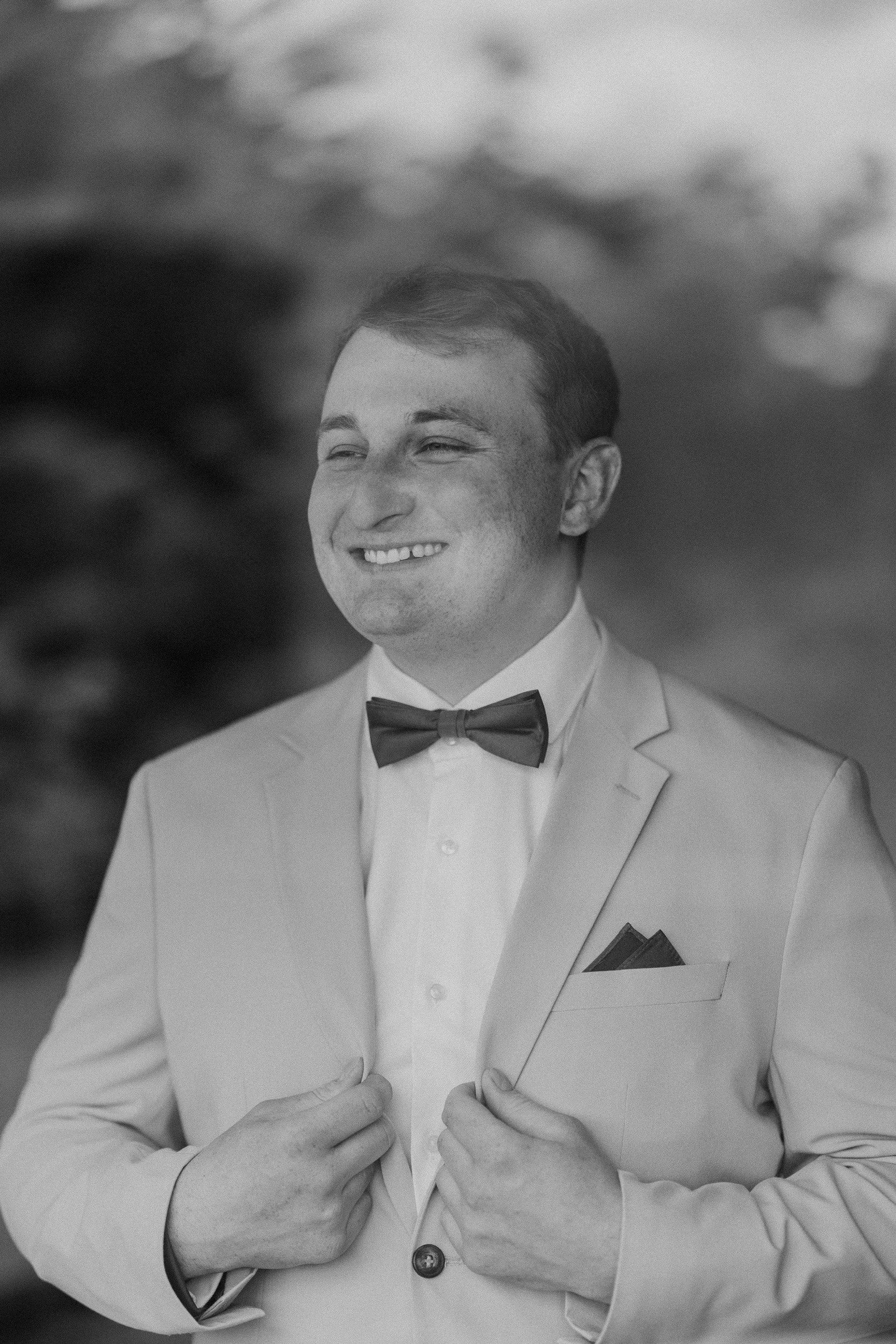 Ein lachender Mann in formeller Kleidung, trägt einen schwarzen Fliege, Anzug und weißes Hemd, steht draußen.