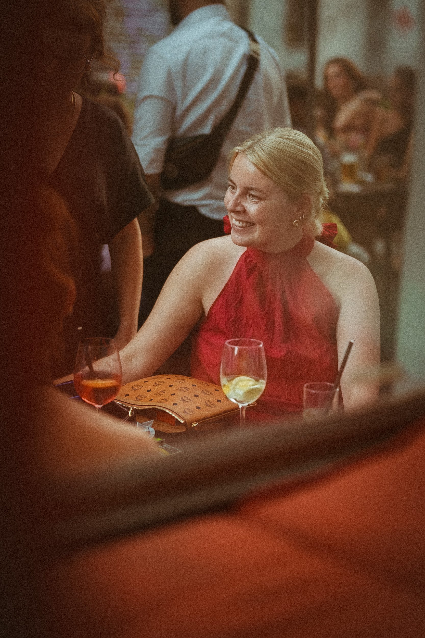 Eine lachende Frau in einem roten Kleid sitzt in einem Restaurant oder einer Bar, umgeben von anderen Personen, mit Getränken auf dem Tisch.