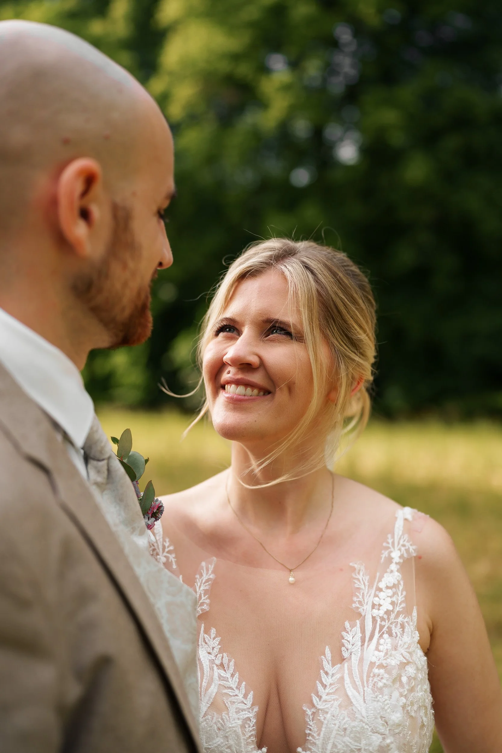 Ein Brautpaar im Freien, die Braut lächelt den Bräutigam an, der Blick ist auf sie gerichtet, im Hintergrund sind Bäume und grünes Laub zu sehen.