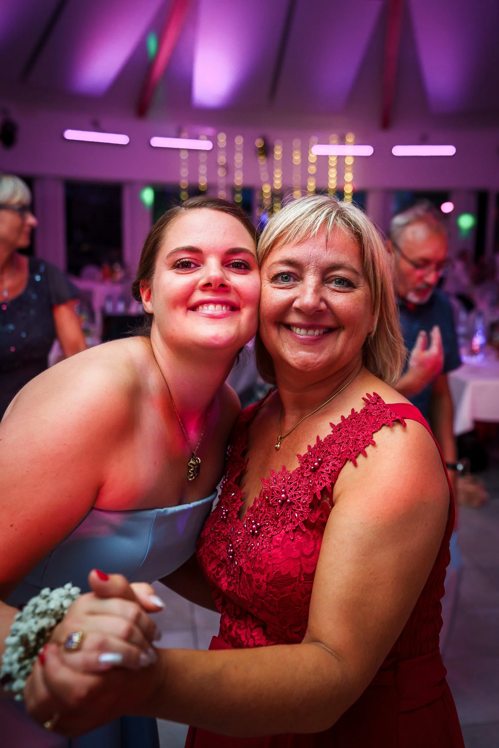 Zwei lachende Frauen tanzen auf einer Feier. Im Hintergrund sind weitere Gäste und bunte Lichter zu sehen.