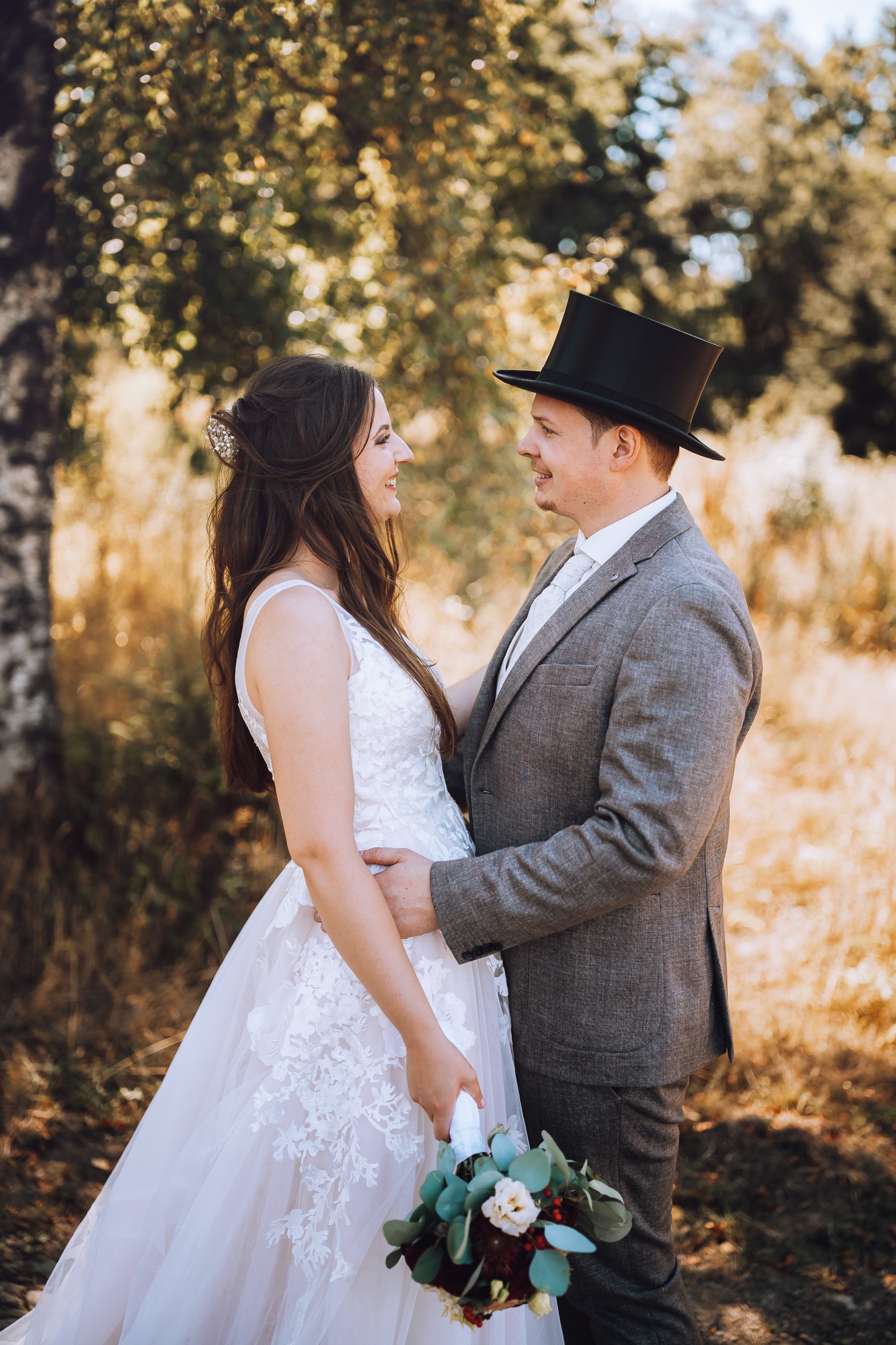 Ein Brautpaar in einem Wald bei Sonnenuntergang, das sich liebevoll ansieht. Der Bräutigam trägt einen grauen Anzug mit Zylinder und die Braut ein weißes Hochzeitskleid, sie hält einen Blumenstrauß.