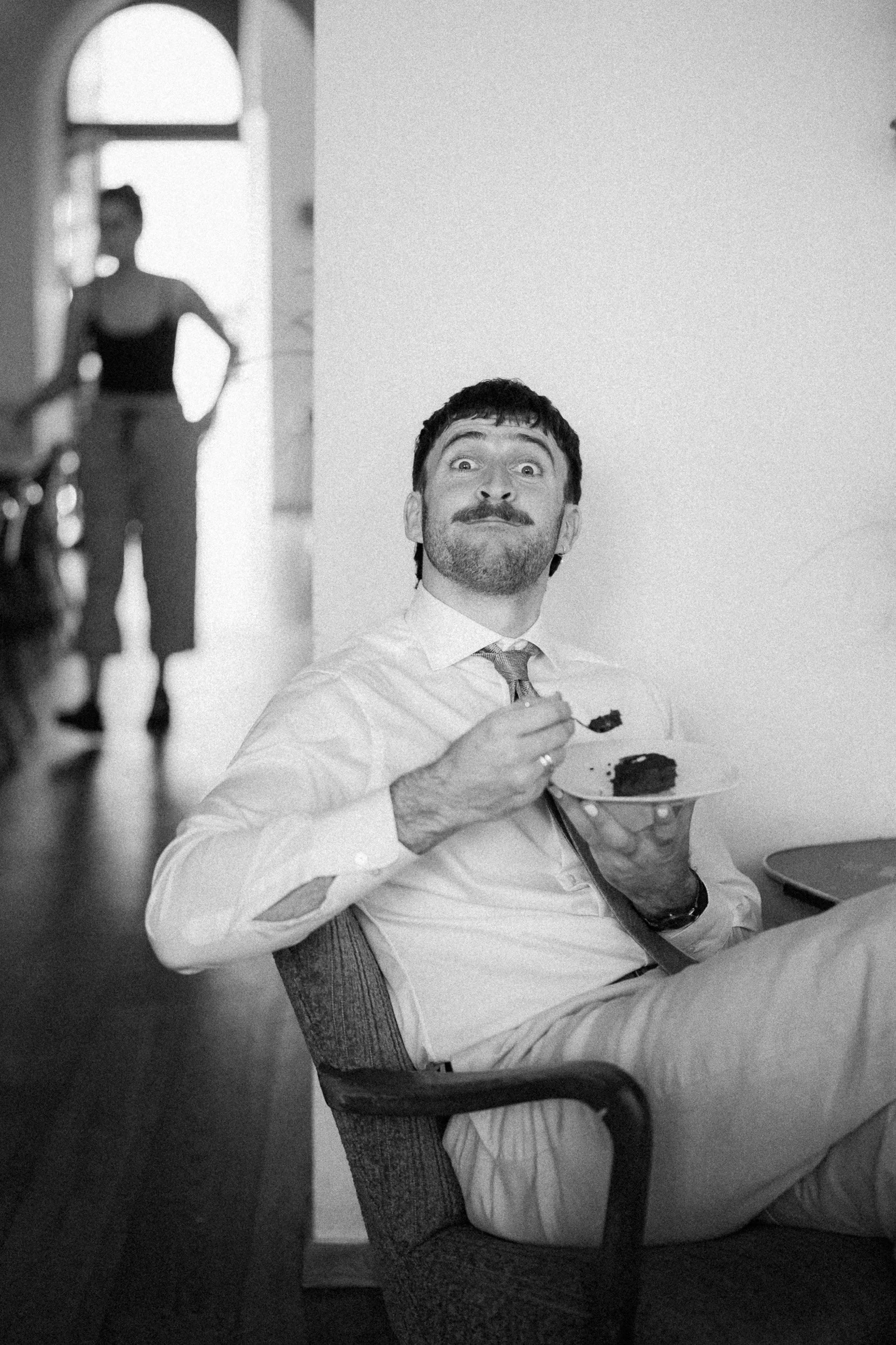 Ein Mann in formeller Kleidung, der auf einem Stuhl sitzt und Kuchen isst, während eine Frau im Hintergrund im Flur steht.