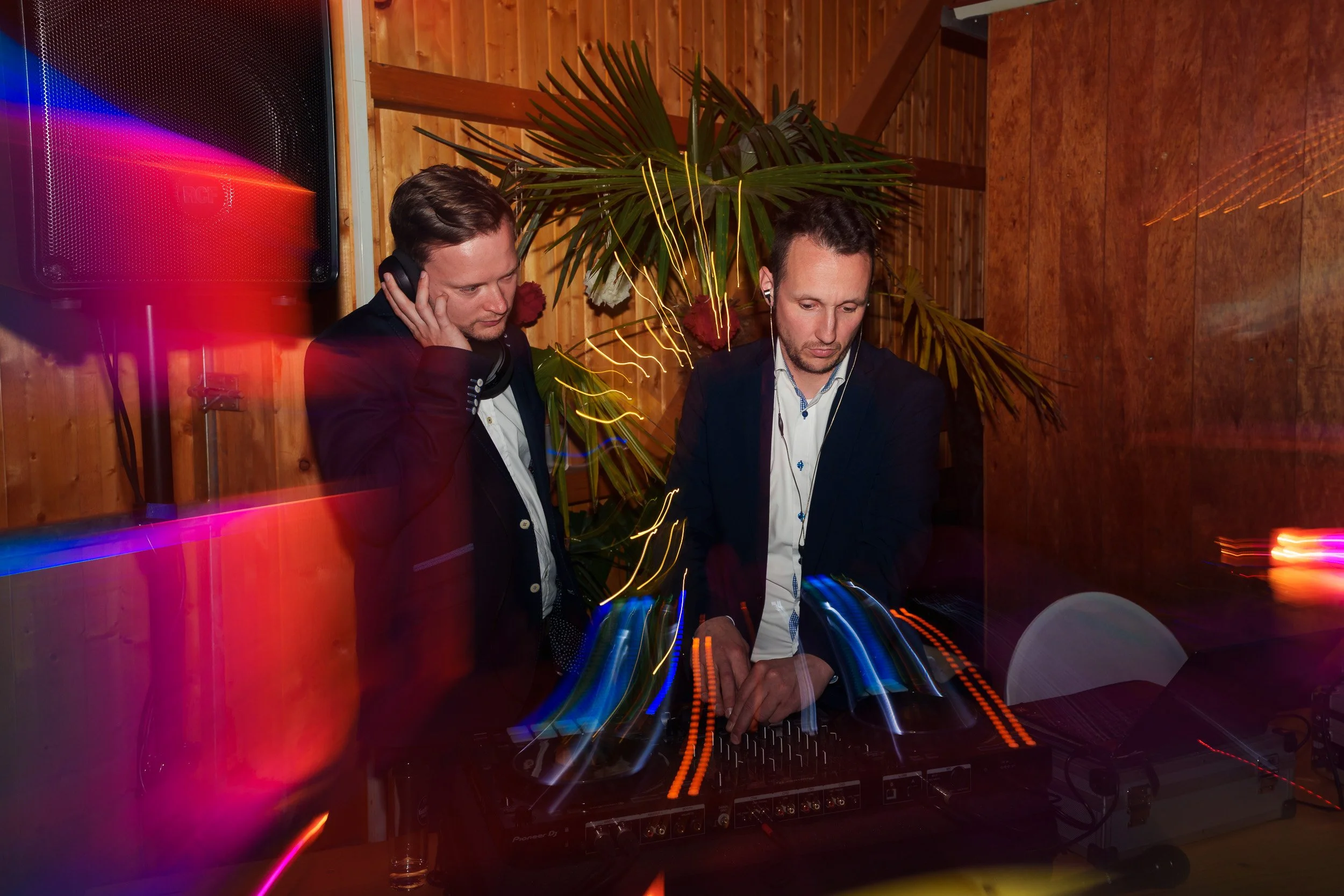 Zwei Männer beim Auflegen in einem Raum mit Holzverkleidung und Pflanzen, bunte Lichter und Bewegungseffekte, die DJ-Ausrüstung.
