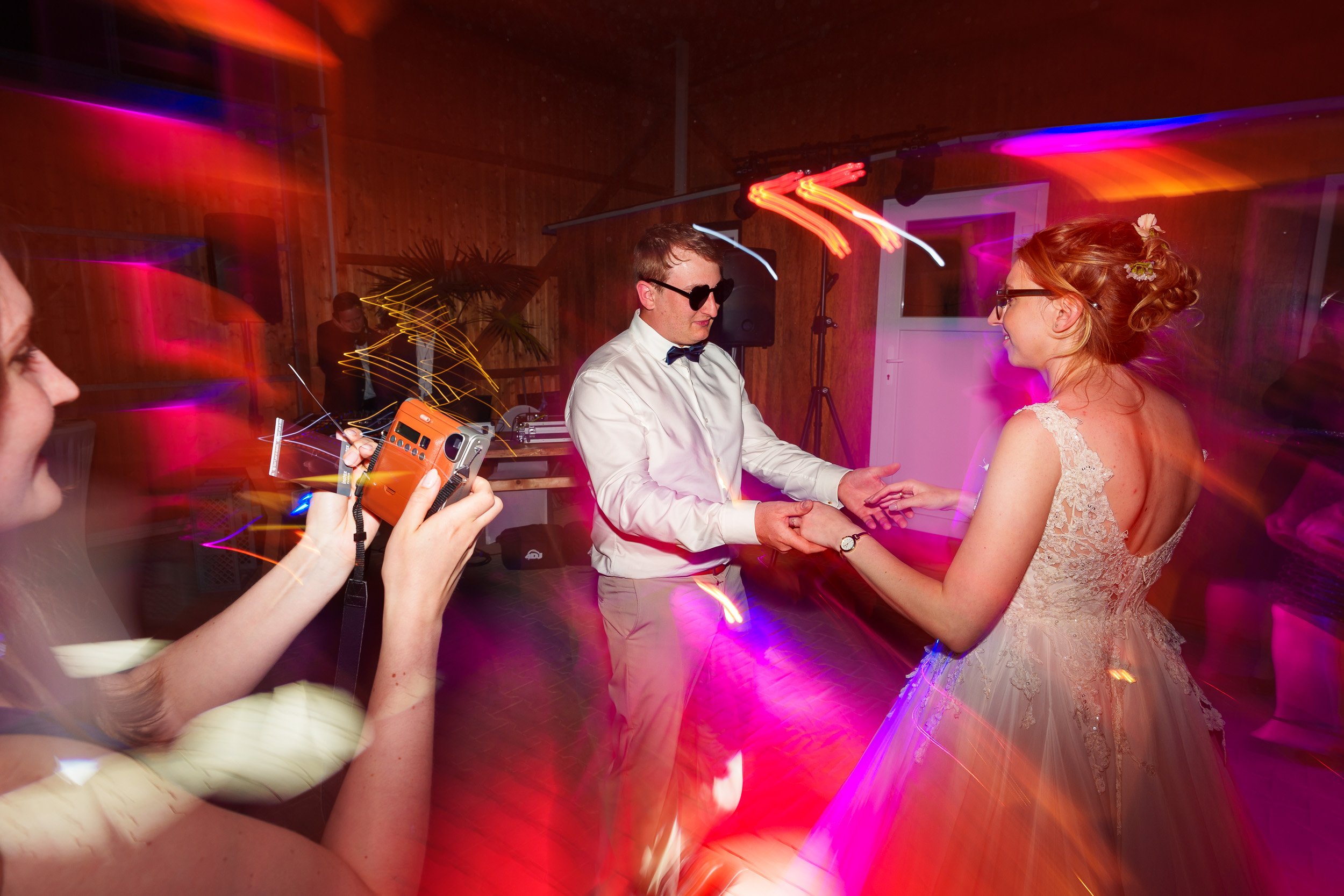 Paare beim Tanzen auf einer Party oder Hochzeit in einem Raum mit Holzwänden, bunte Lichter, eine Frau filmt mit einer Kamera, ein Mann in Sonnenbrille und Fliege tanzt mit einer Frau in einem weißen Hochzeitskleid.