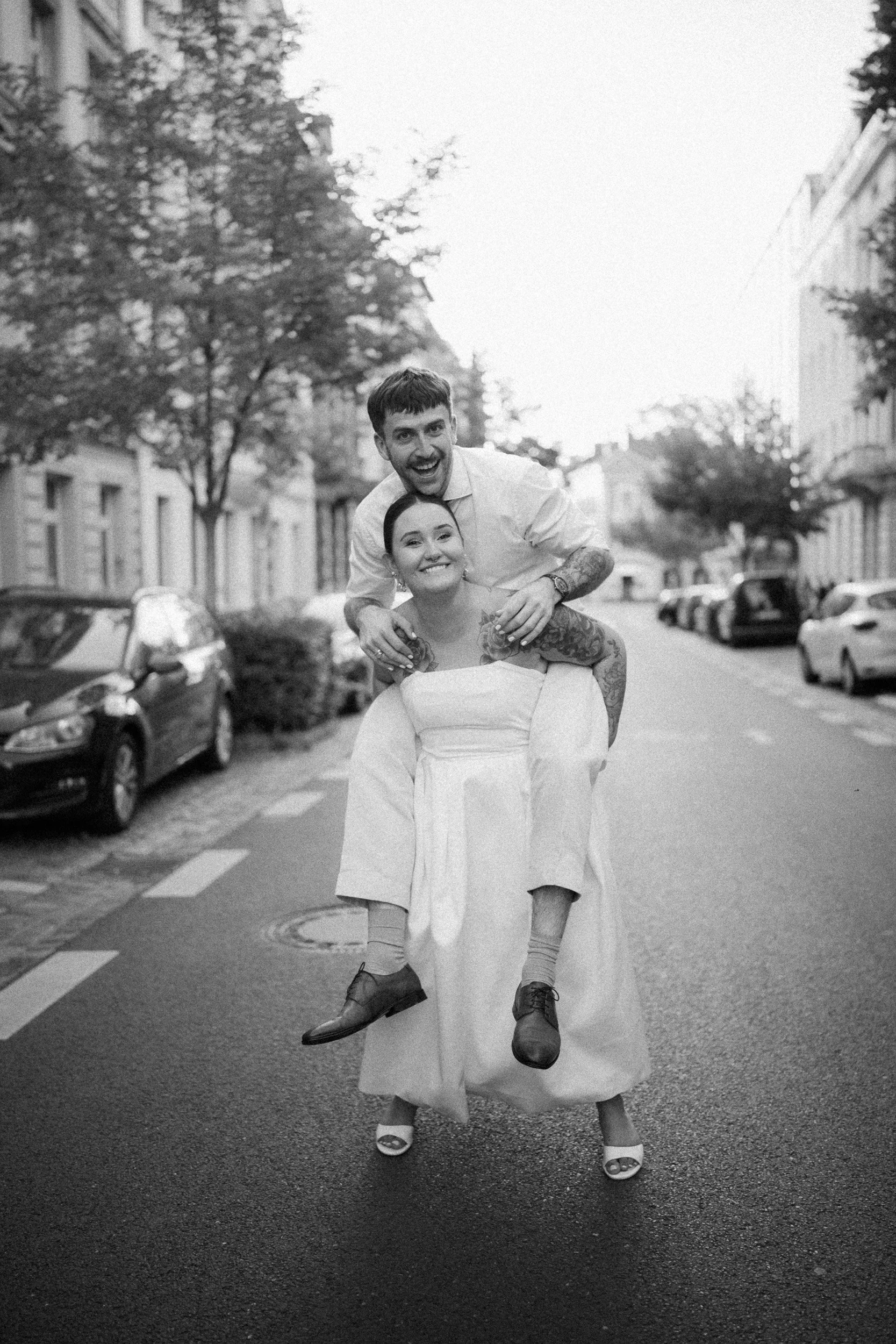 Ein Paar in formeller Kleidung, auf einer Stadtstraße, der Mann sitzt auf den Schultern der Frau, beide smilen glücklich, es ist schwarz-weiß Fotografie.