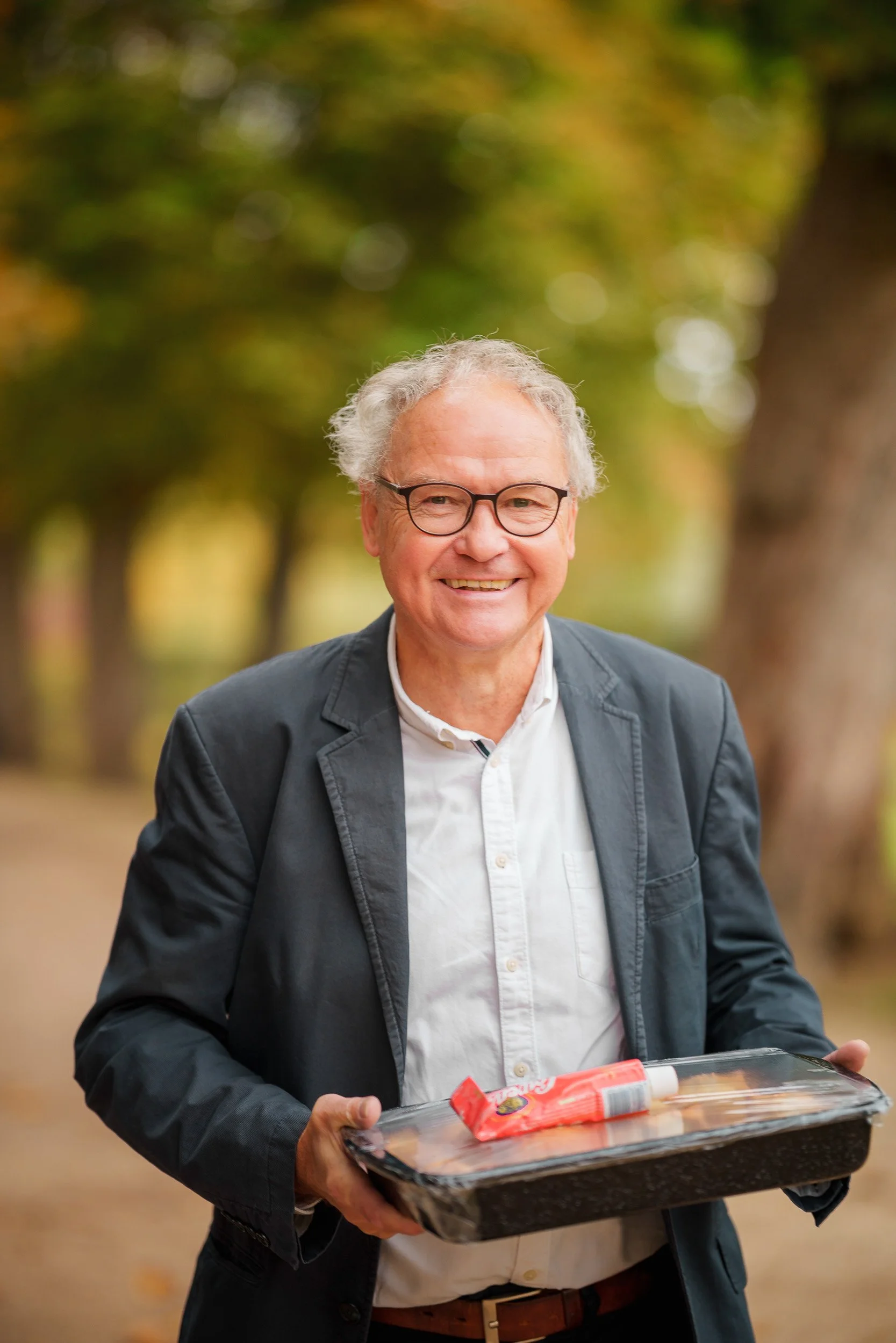 Ein lächelnder älterer Mann mit Brille, trägt einen dunklen Blazer und ein weißes Hemd, hält einen mit Lebensmittel verpackten Behälter in der Hand, im Hintergrund Bäume mit herbstlichen Blättern.
