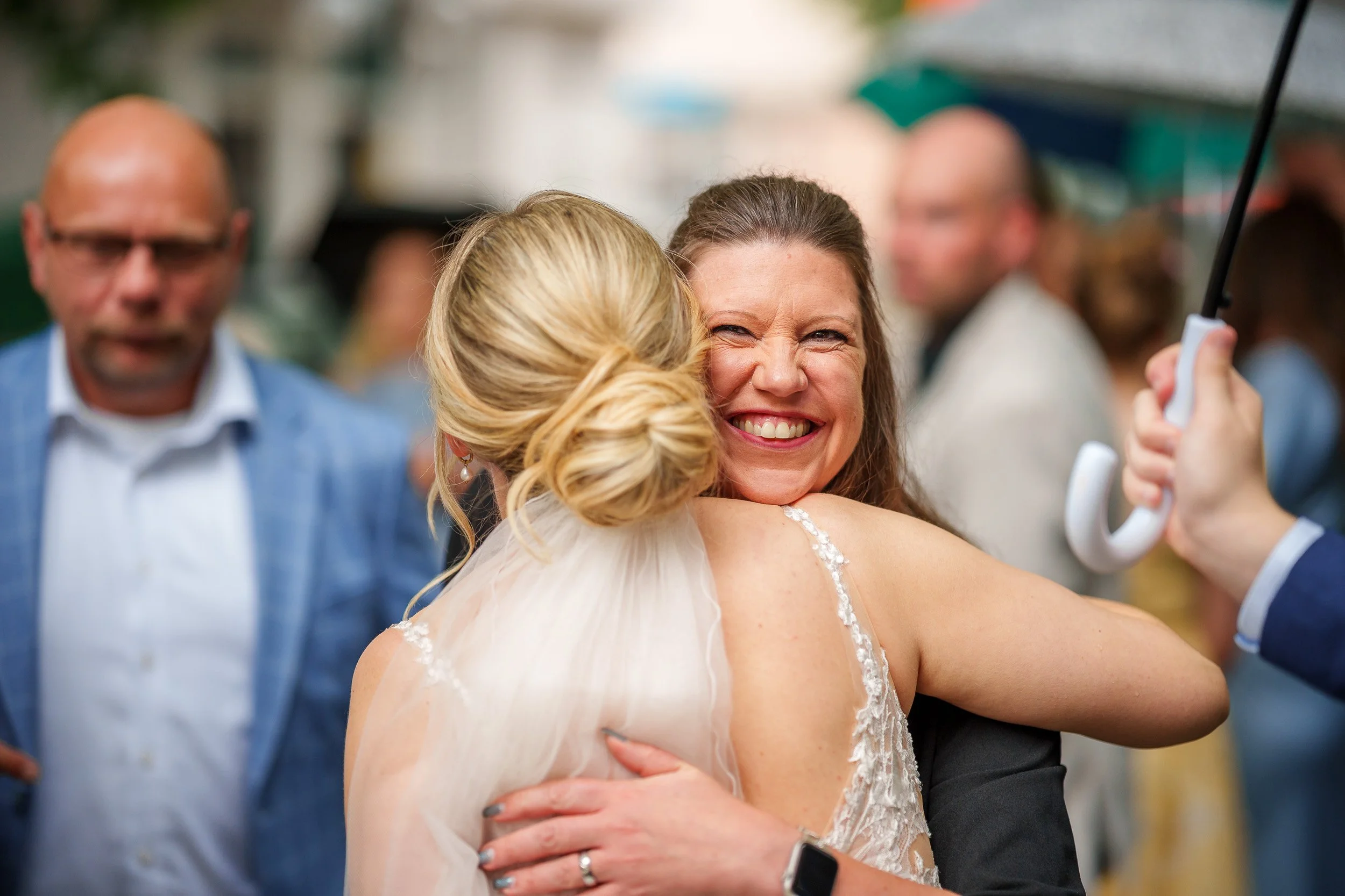 Zwei Frauen umarmen sich auf einer Feier, eine lächelt breit, während sie sich freuen, im Hintergrund sind weitere Menschen und Regenschirme sichtbar.