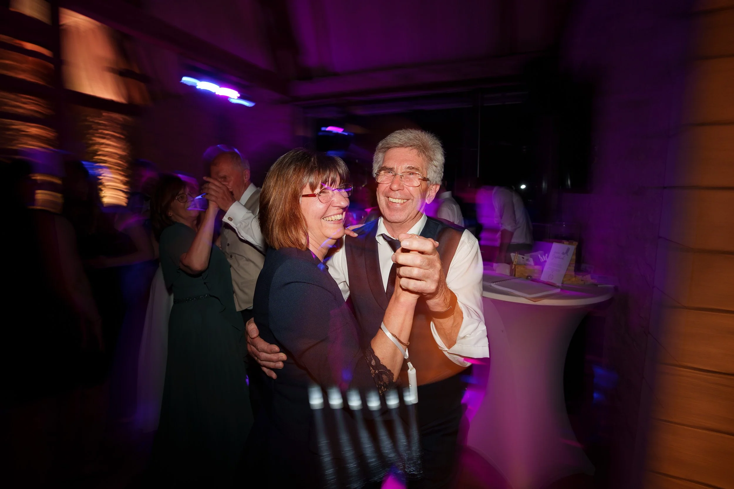 Paar tanzt auf einer Party, umgeben von weiteren Feiernden im Hintergrund, in einem Raum mit dunkler Beleuchtung und bunten Lichtern.