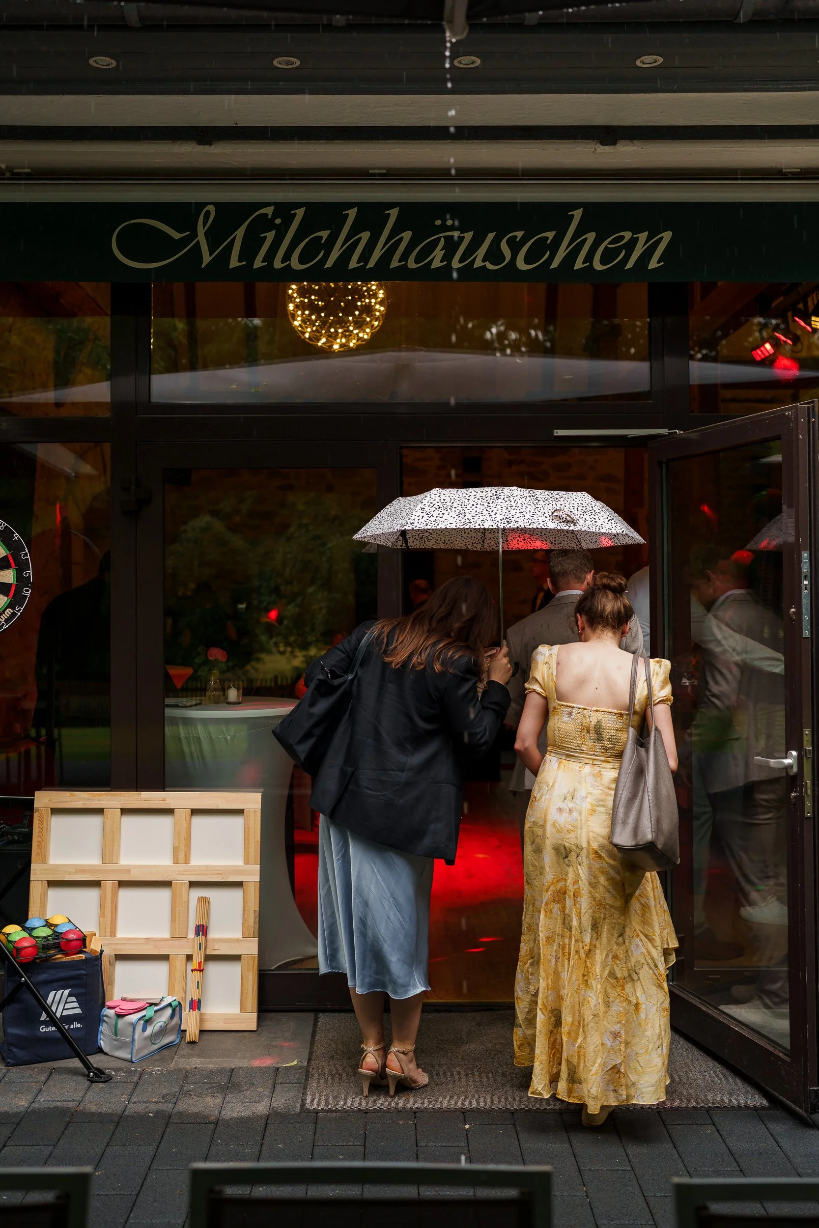 Menschen betreten ein Restaurant namens Milchhäuschen bei Regen, eine Frau hält einen Regenschirm, das Restaurant hat dunkle Türen und eine freundliche Atmosphäre.