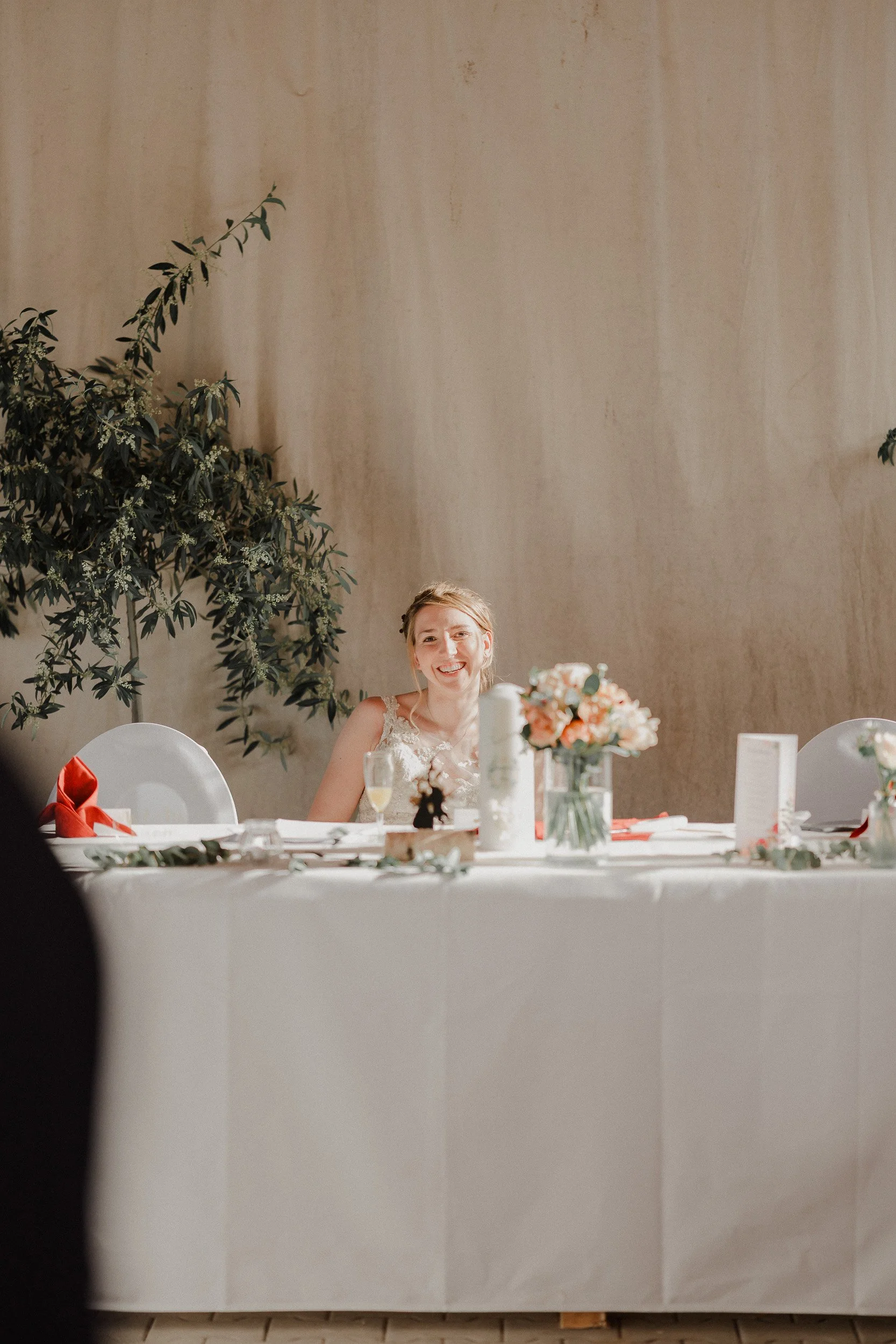 Eine lächelnde Braut sitzt an einem festlich gedeckten Tisch bei einer Hochzeit.