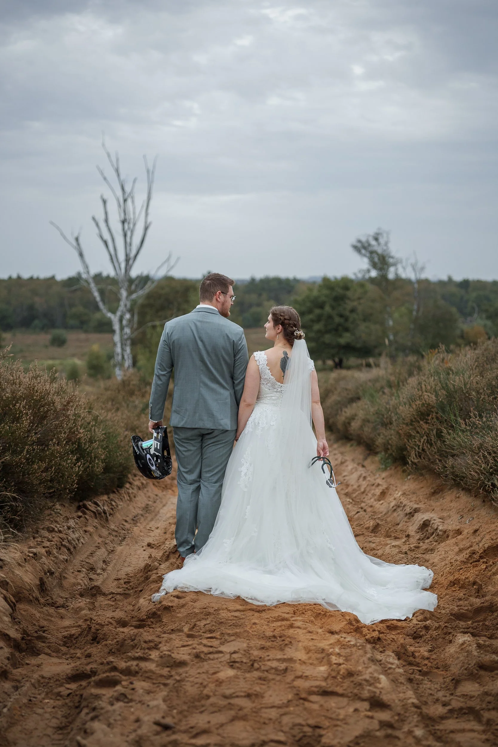 Ein Brautpaar in Hochzeitskleidung läuft auf einer landwirtschaftlichen Straße in der Natur, das Mädchen trägt ein weißes Brautkleid mit Schleier und das Mädchen einen grauen Anzug und hält einen Helm in der Hand.