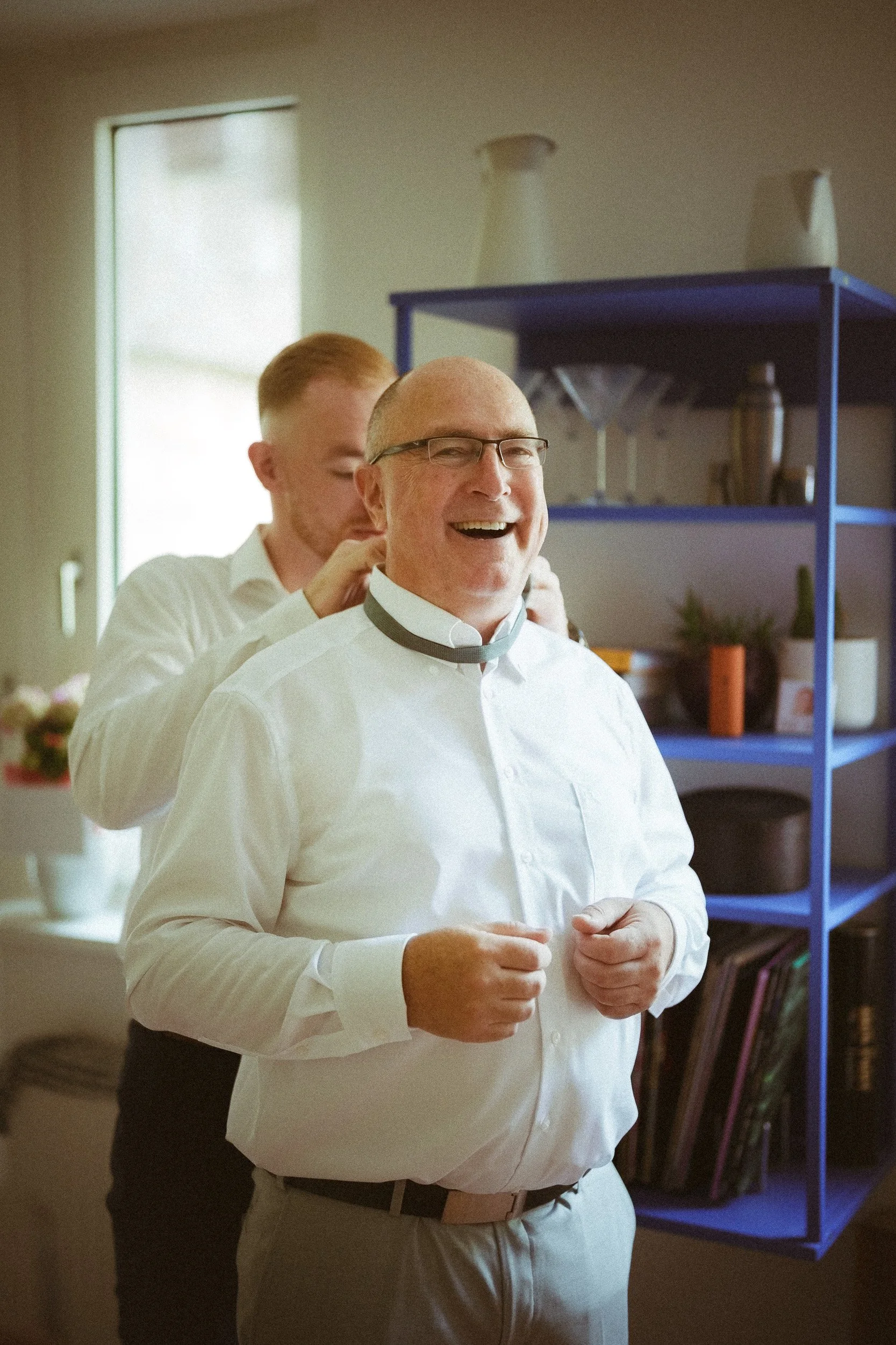 Ein älterer Mann wird von einem jüngeren Mann an seinem Halsgurt beim Anziehen eines Hemdkragens unterstützt, beide lachen im Wohnzimmer.