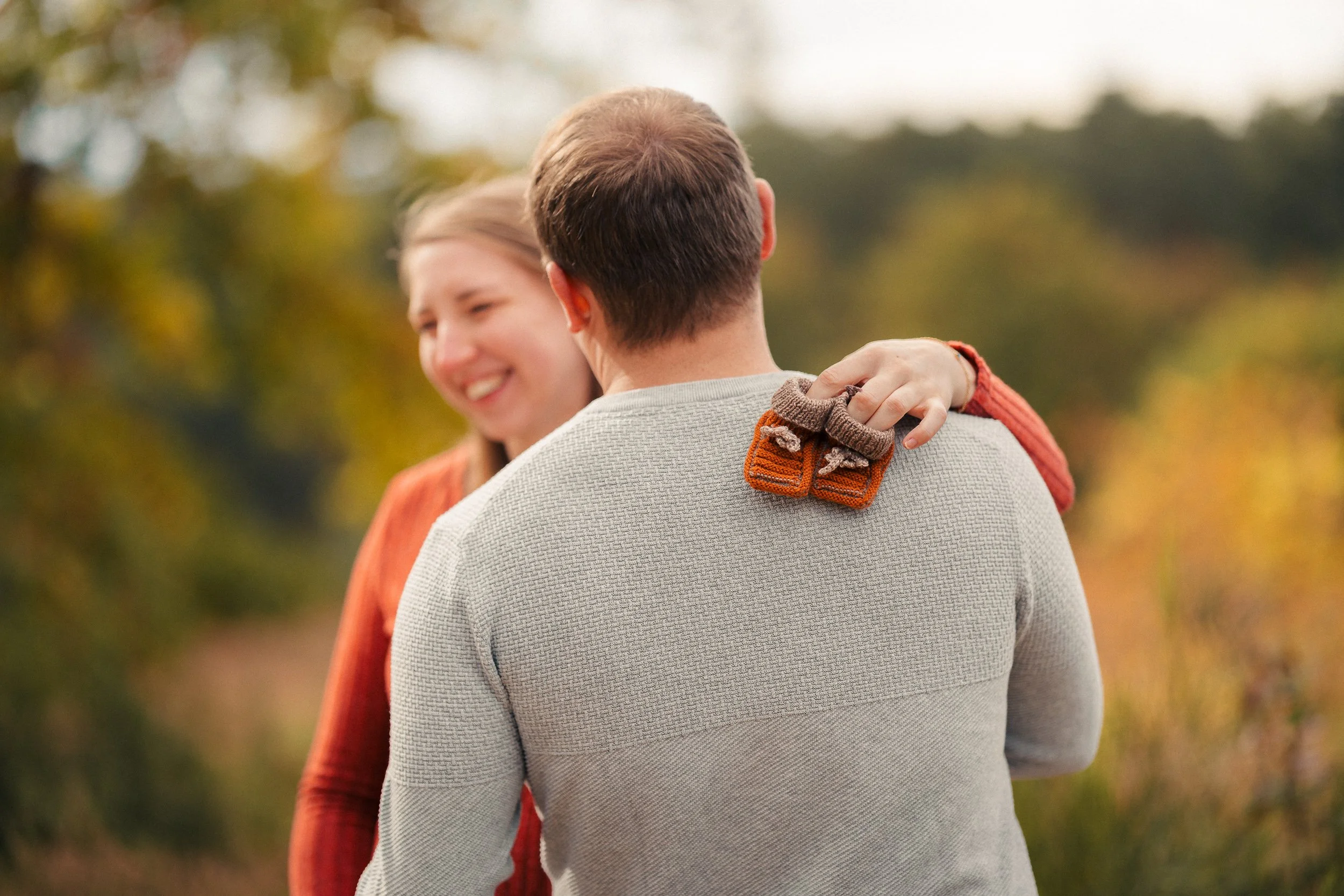 Ein Mann und eine Frau umarmen sich draußen in einer herbstlichen Landschaft, die Frau lächelt glücklich.