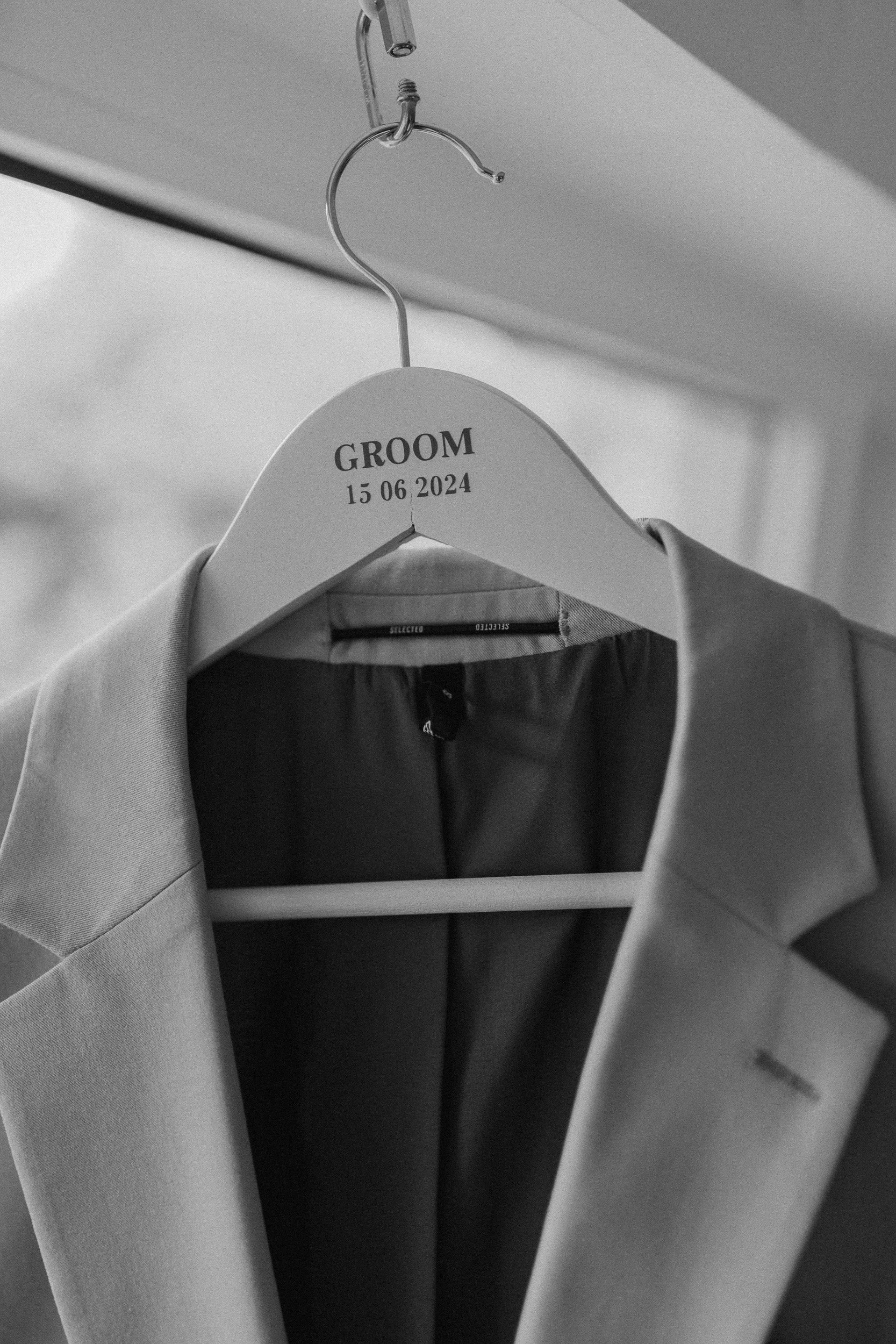 Ein schwarzer und weißer Sakko hängt an einem Kleiderbügel, der mit der Aufschrift 'GROOM' und dem Datum '15 06 2024' versehen ist.