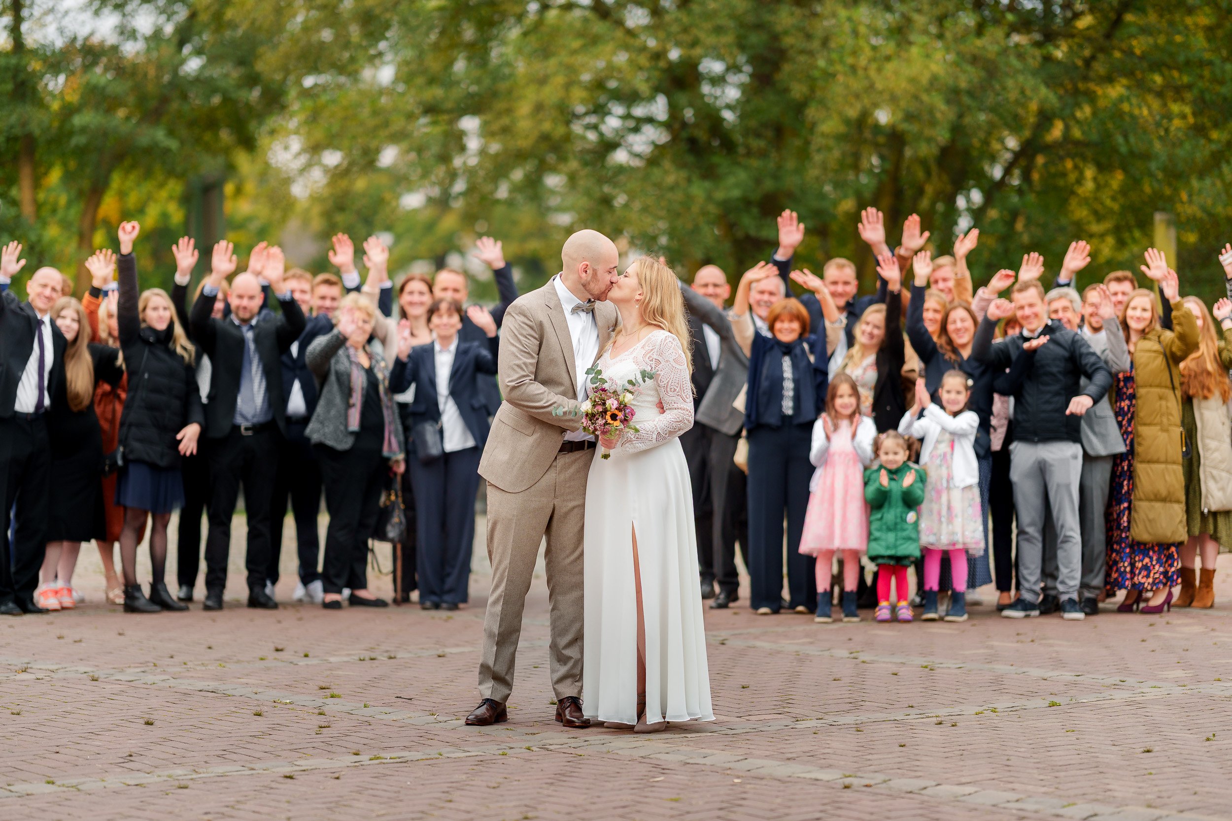 Ein Brautpaar küsst sich, umgeben von einer Gruppe von stehenden Hochzeitsgästen im Park.