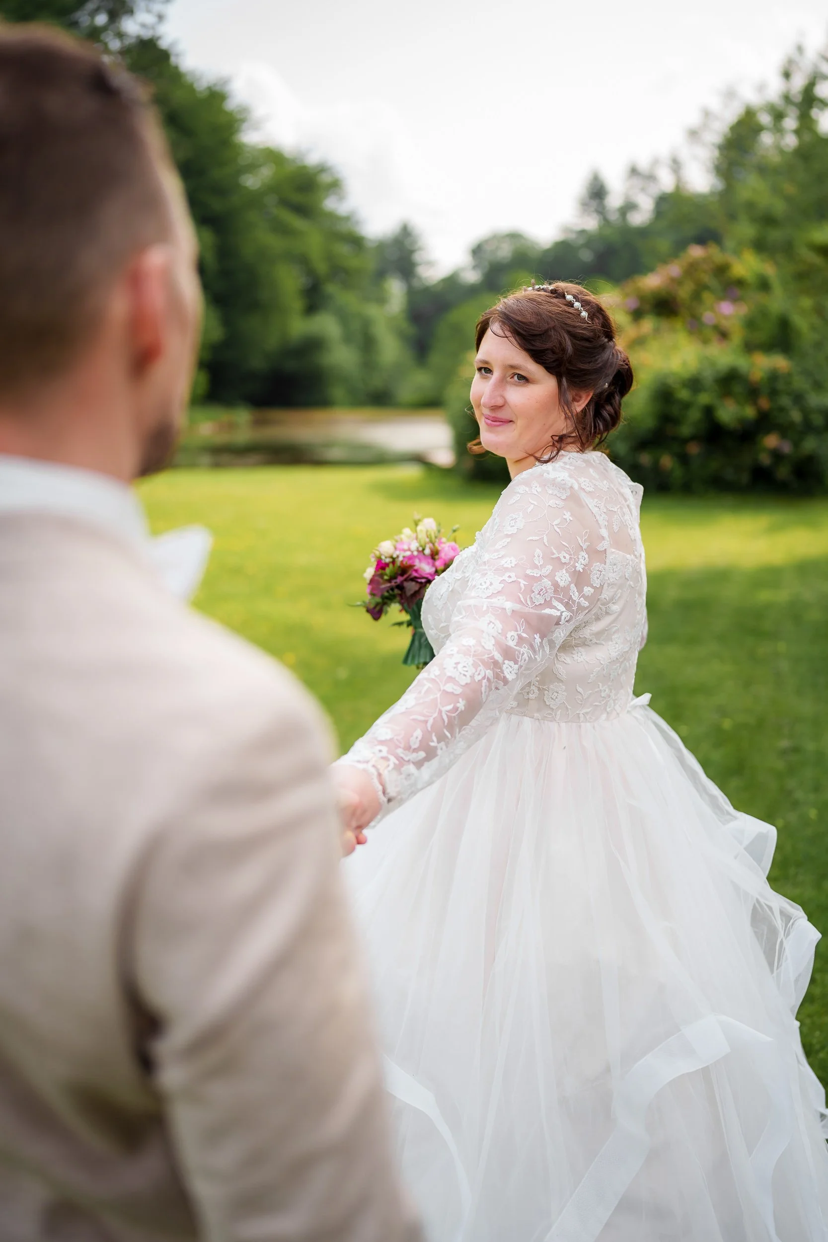 Braut hält die Hand des Bräutigams, auf einer grünen Wiese bei Diana im Park, im Vordergrund ist der Bräutigam unscharf, im Hintergrund sieht man Bäume und einen Wasserlauf.