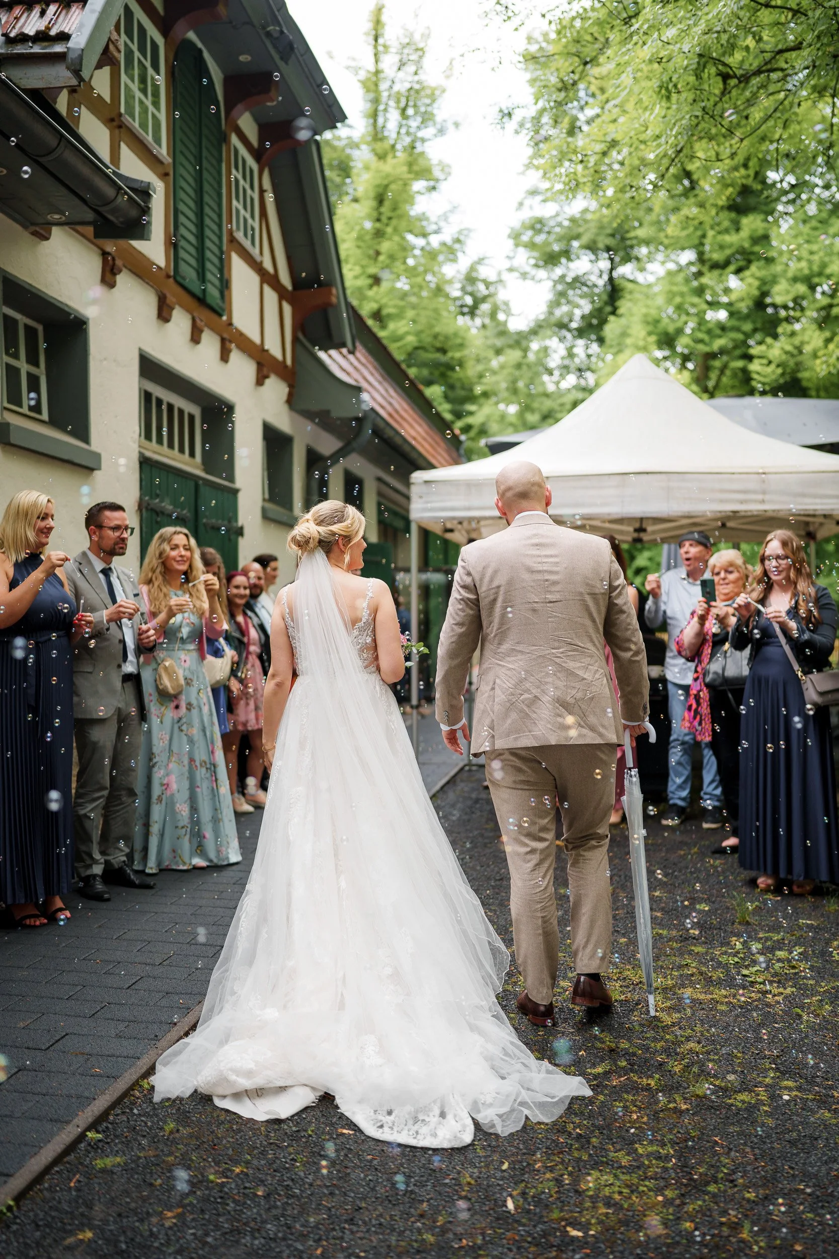 Braut und Bräutigam gehen nach ihrer Hochzeit an einer Feier vorbei, umgeben von Freunden und Familie, in einer grünen, bewaldeten Umgebung.