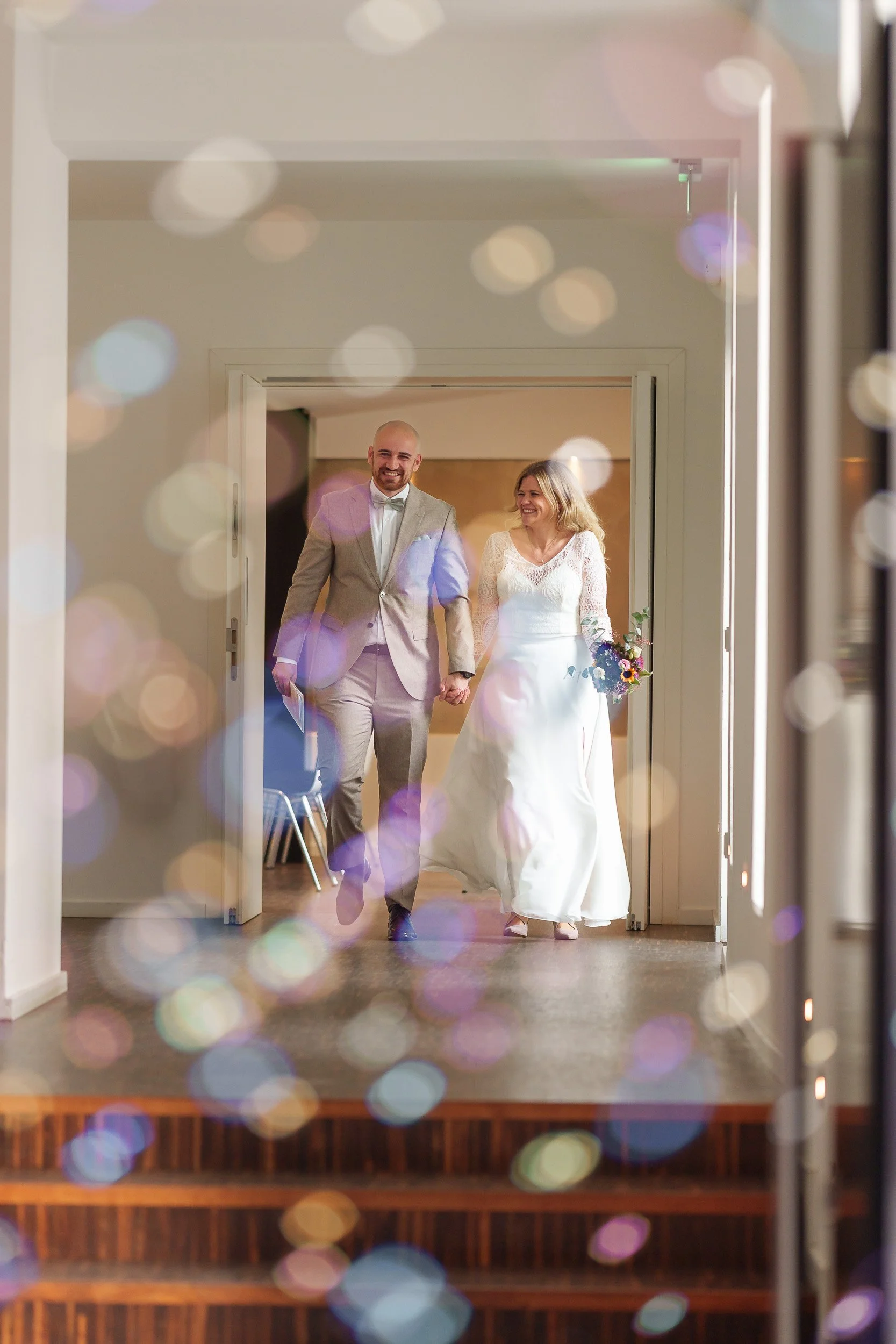 Ein frisch verheiratetes Paar in formeller Kleidung hält Händchen und läuft glücklich durch einen Raum. Die Szene ist mit bunten Lichtreflexen und Bokeh-Effekten dekoriert.