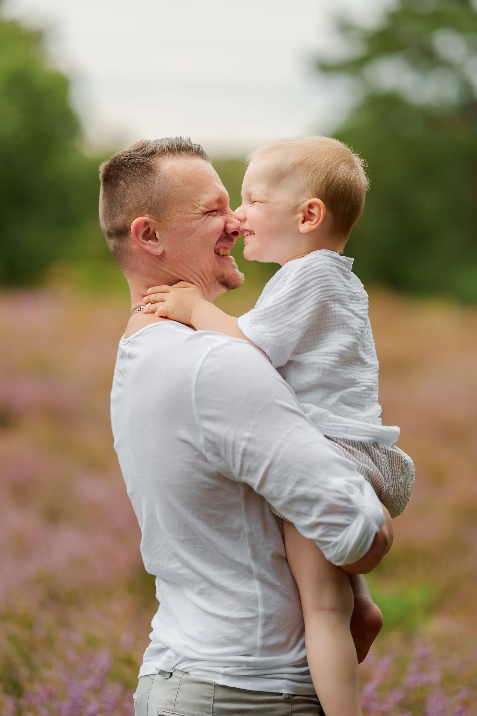 Vater hält seinen lachenden kleinen Sohn im Freien, im Hintergrund eine Blumenwiese mit pinken Blüten, Umgebungsbäume und Himmel, beide tragen weiße Kleidung.