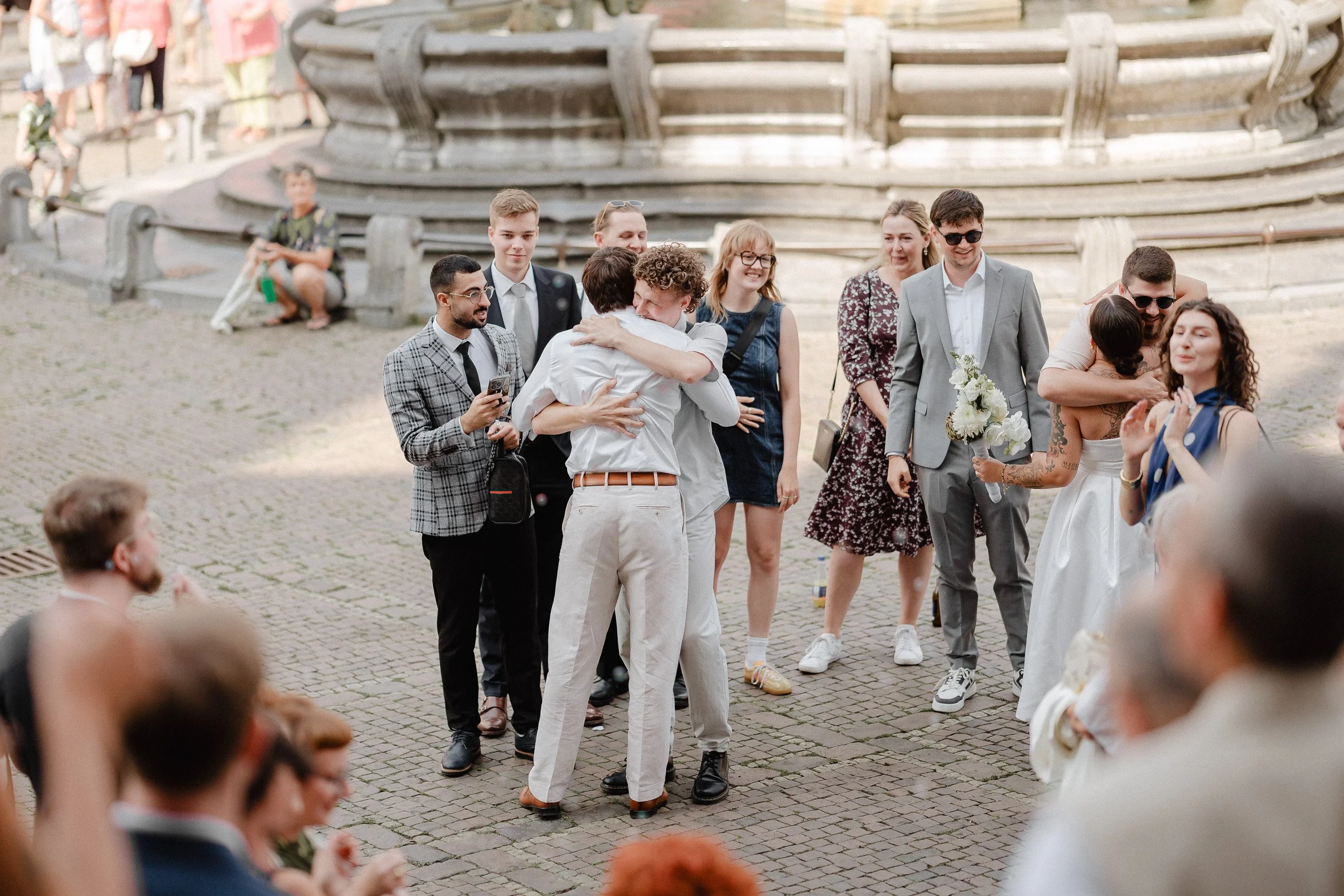 Hochzeitsszene mit Brautpaar, Freunden und Gästen im Freien auf einem gepflasterten Platz, umgeben von einer historischen Statuen- oder Brunnenstruktur.