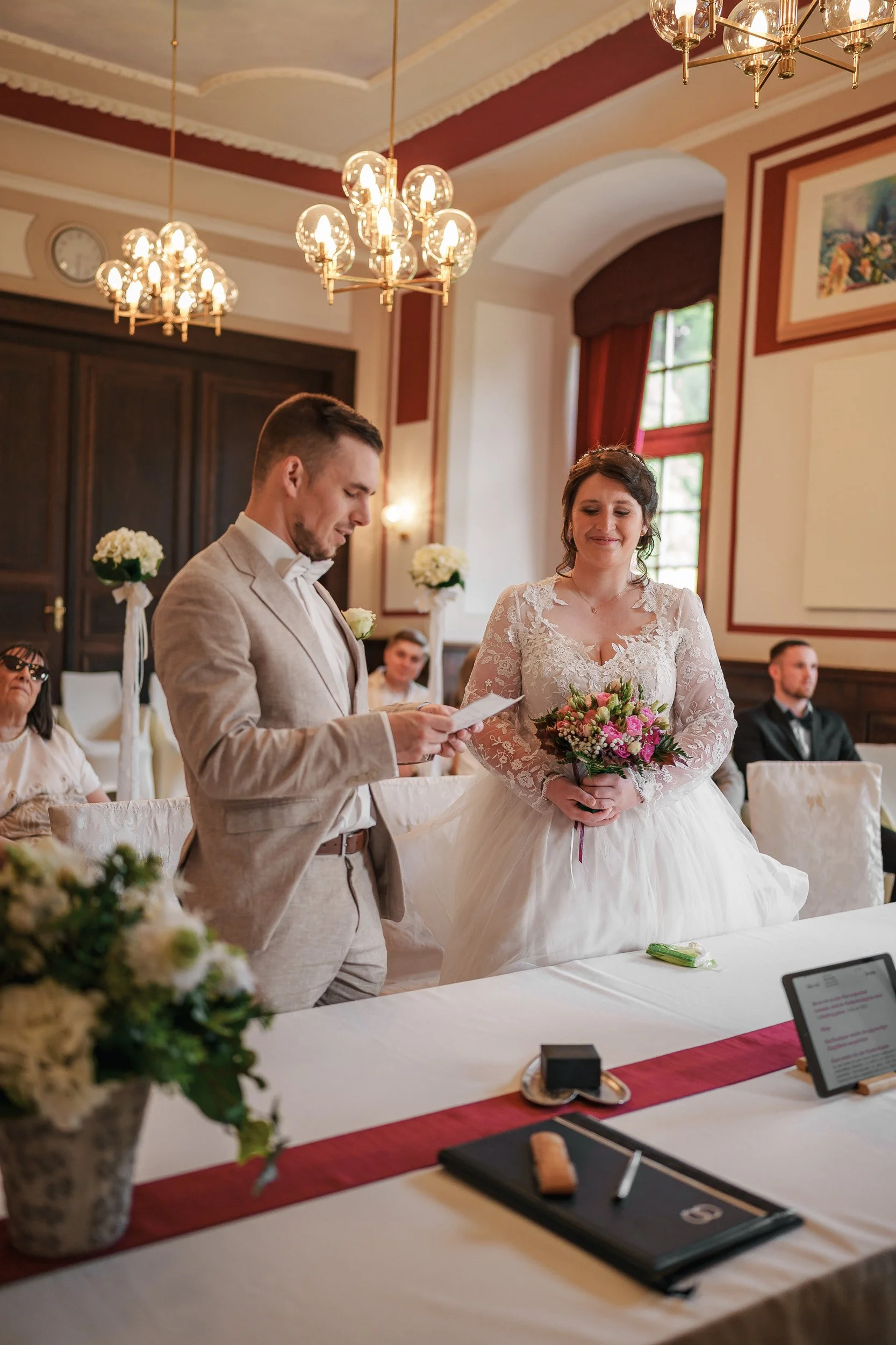 Braut und Bräutigam während ihrer Trauung in einem festlich dekorierten Raum, mit Gästen im Hintergrund, Blumen und Kerzen auf dem Tisch