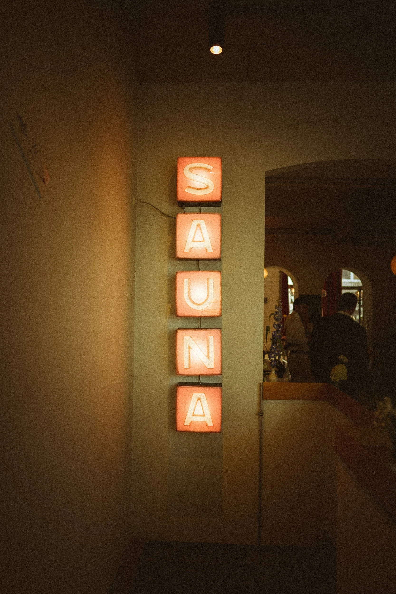 Lichtreklame mit vertikaler Anordnung der Buchstaben "SAUNA" an der Wand in einem Raum mit gedämpftem Licht, im Hintergrund sind Menschen und ein Fenster sichtbar.
