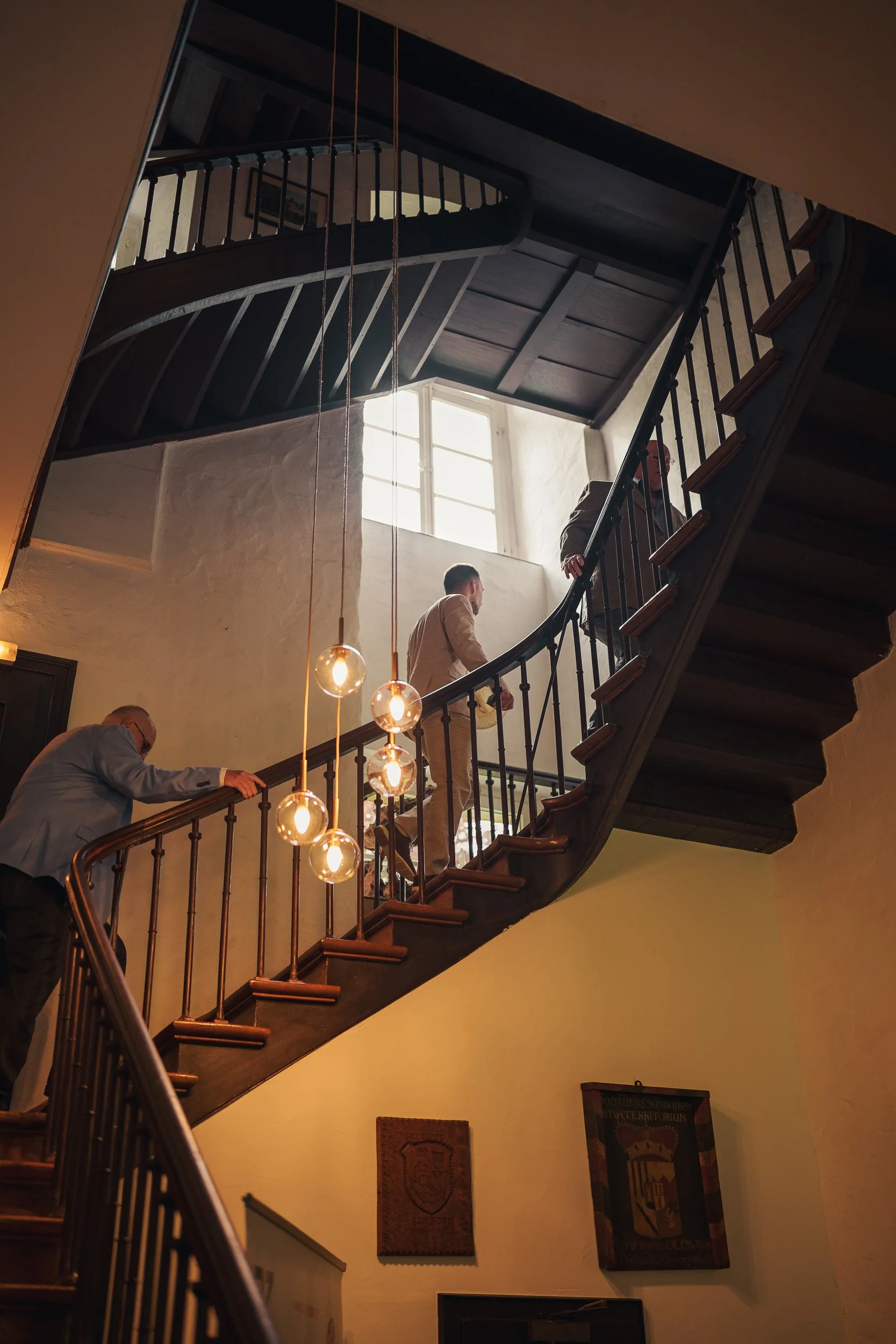 Drei Menschen gehen eine Treppe in einem alten Gebäude hinauf, mit großen Fenstern und einer modernen Beleuchtung mit Glühbirnen.