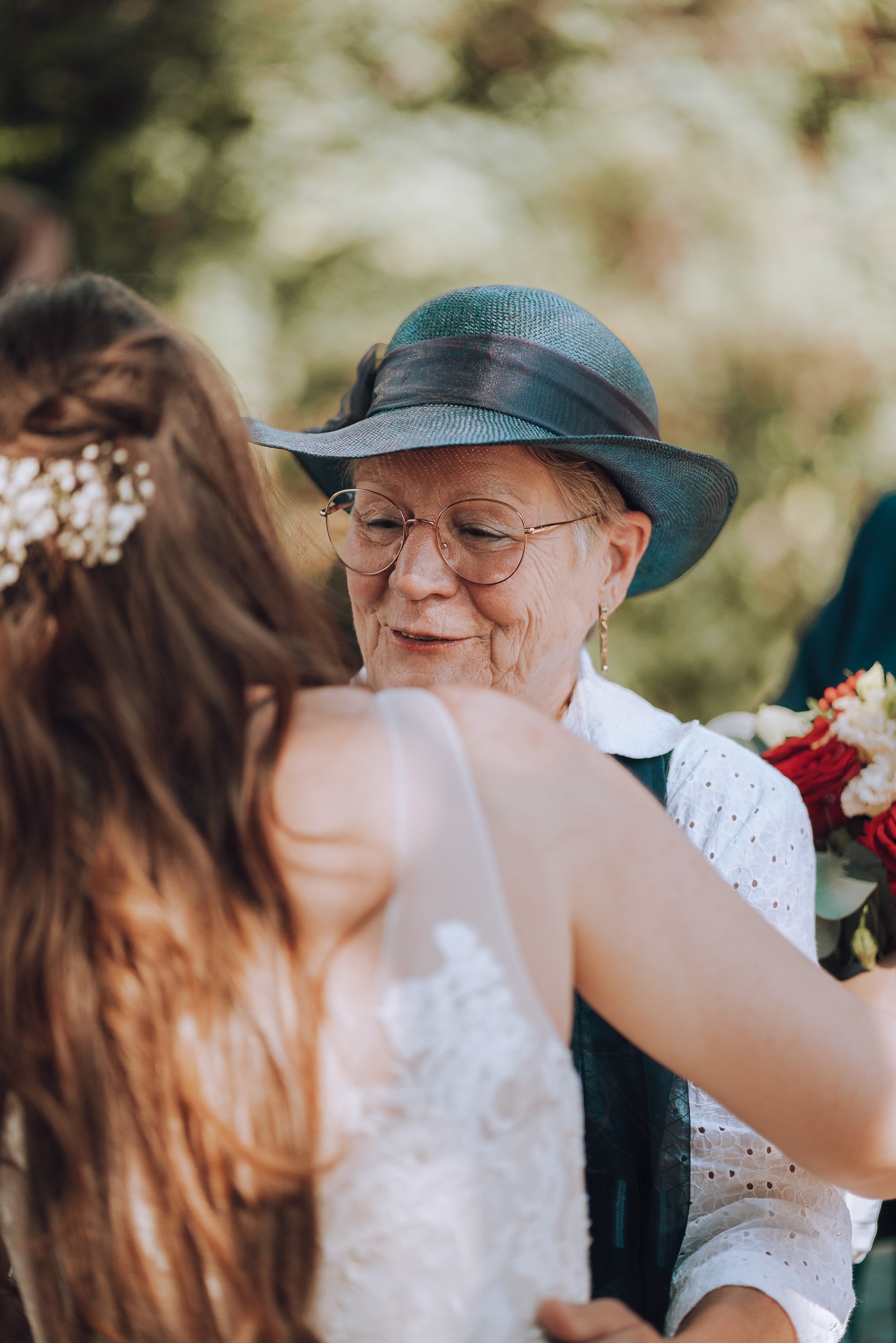 Eine ältere Frau trägt eine blaue Hut und Brille, sie umarmt ein junges Mädchen bei einer outdoor Veranstaltung, wahrscheinlich eine Hochzeit, mit Blumen im Hintergrund.