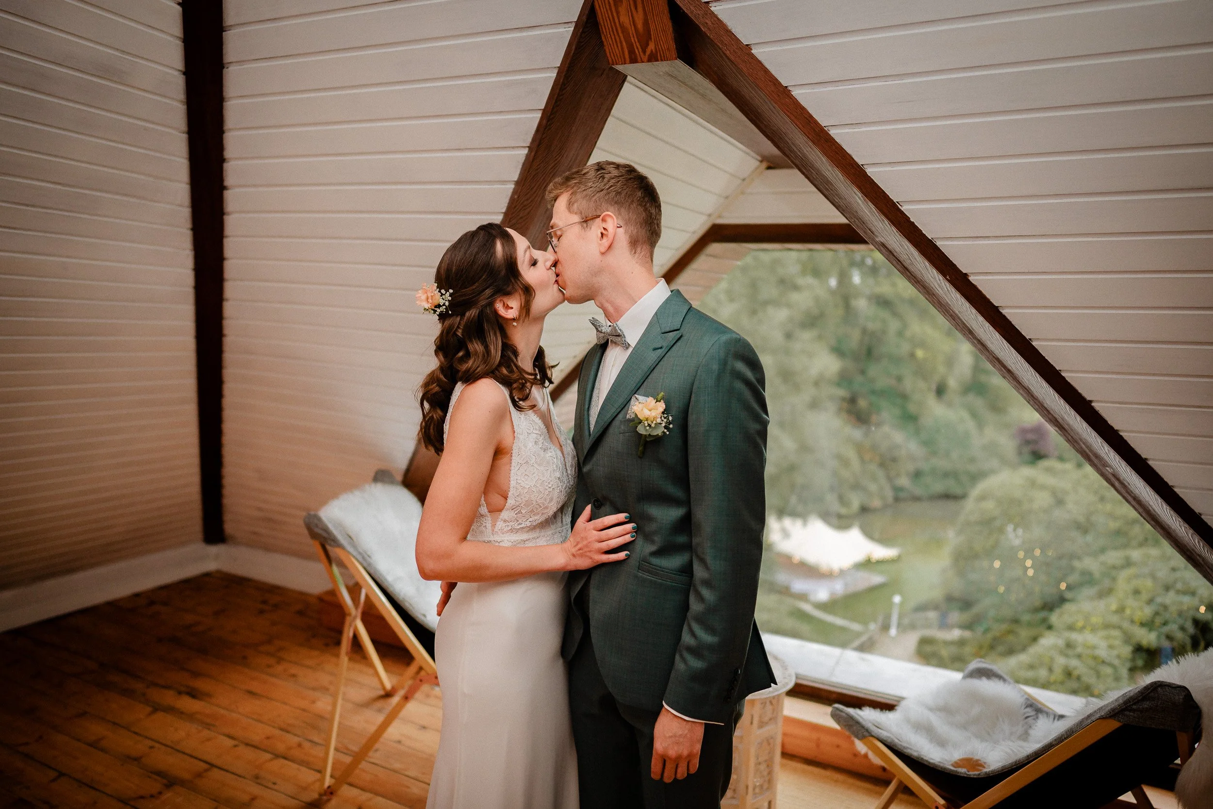Ein brautkleid-tragendes weibliches Paar küsst sich in einem Raum mit Dachschräge und großem Fenster, im Hintergrund eine grüne Landschaft.