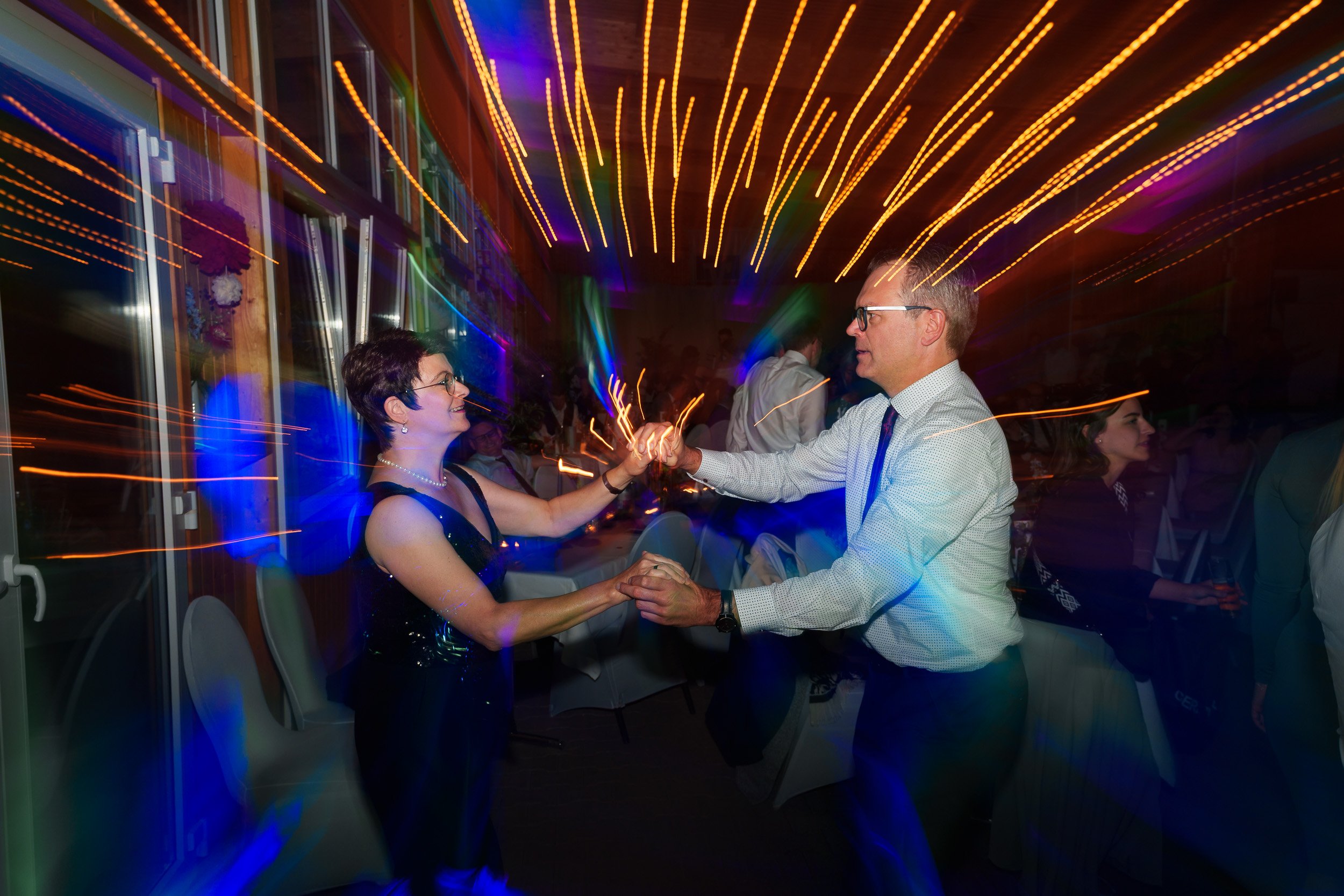 Ein Mann und eine Frau tanzen sich bei einer Feier im Raum, umgeben von bunten Lichtern, während andere Gäste im Hintergrund sitzen und sich unterhalten.