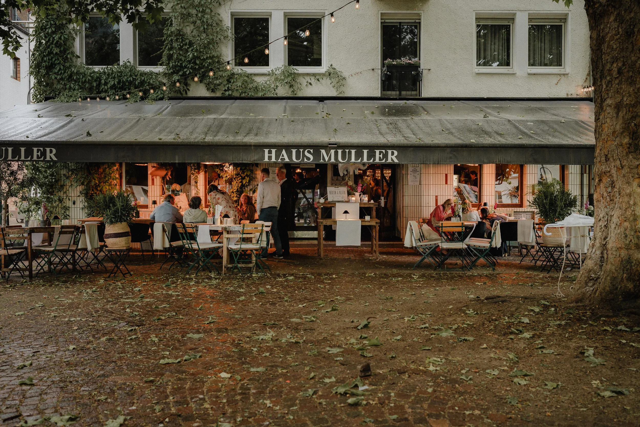 Außenaufnahme eines Restaurants namens haus muller mit Tischen und Stühlen im Freien, umgeben von Bäumen und fallenblättern, bei Dämmerung