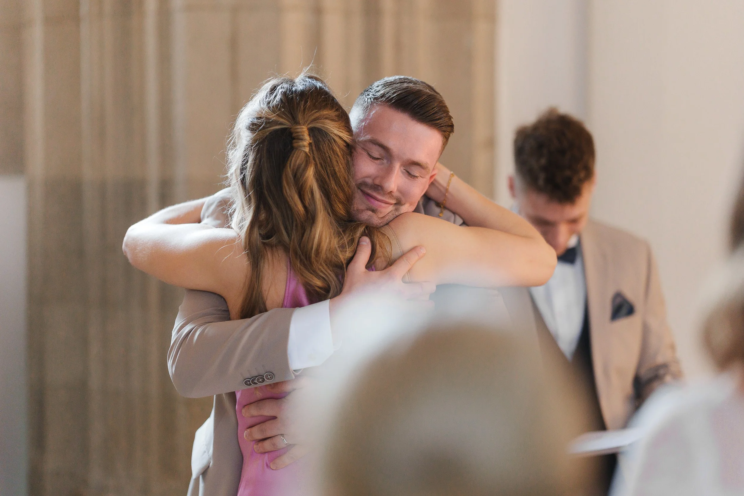 Ein Mann und eine Frau umarmen sich in einem Raum, während im Hintergrund ein weiterer Mann sitzt.