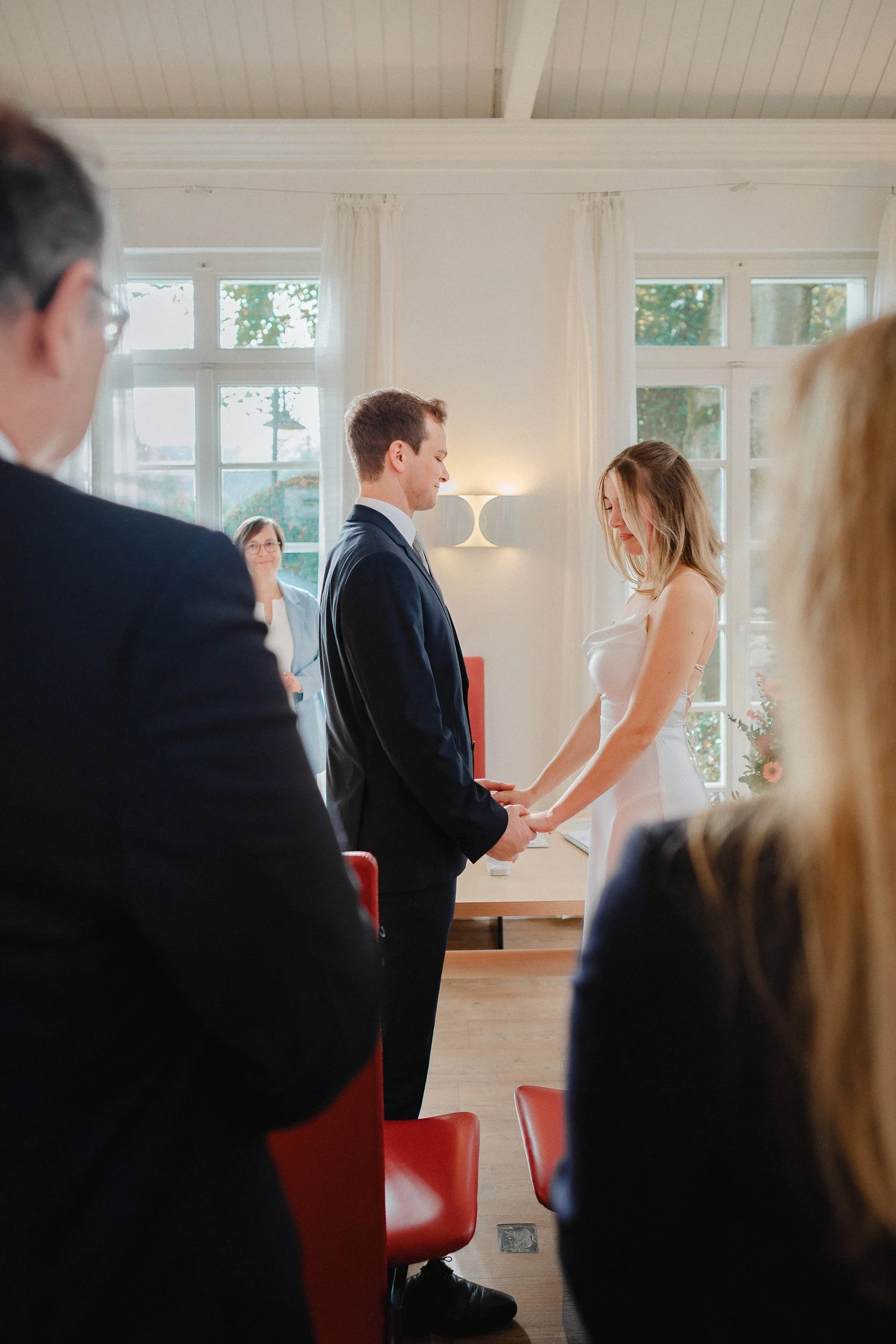 Ein Brautpaar hält sich während einer Trauung im Standesamt bei der Hand, im Hintergrund steht eine Frau in einem blauen Anzug.