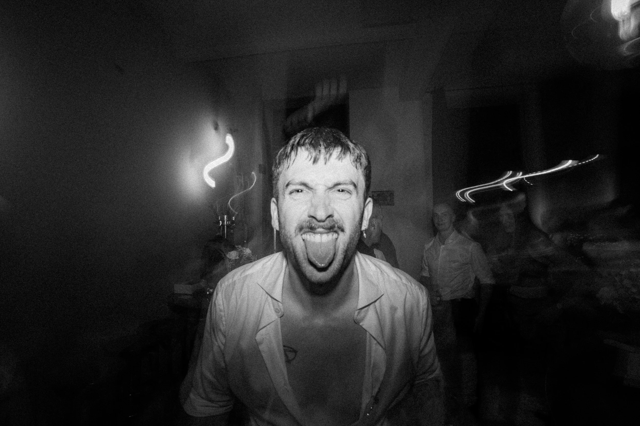 Junge Mann in schwarzem und weißem Hemd zeigt die Zunge in einer dunklen, lebhaften Partyumgebung.