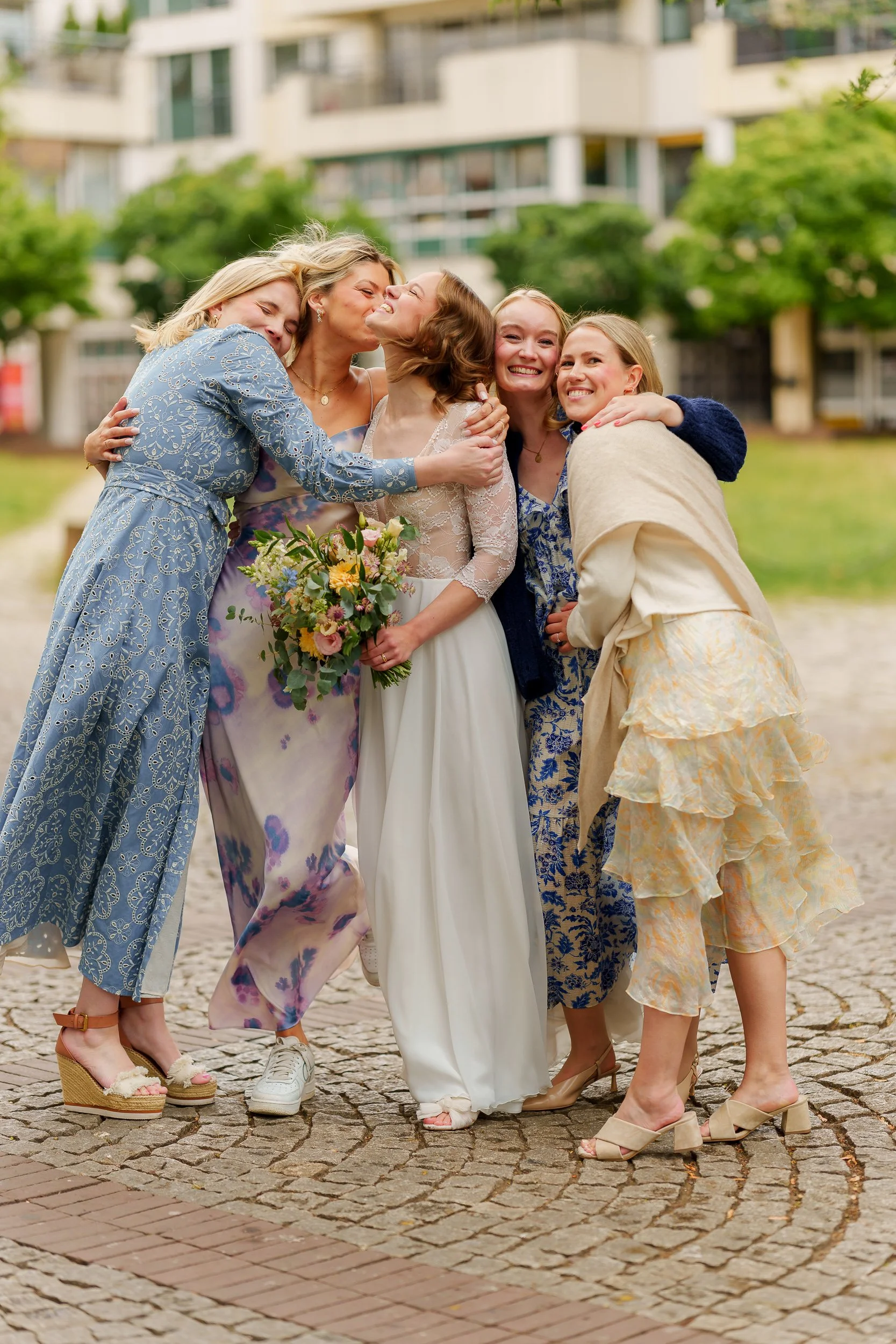 Eine Gruppe von fünf Frauen bei einer Hochzeitsfeier, die sich herzlich umarmen und lachen, im Freien vor einem modernen Gebäude.