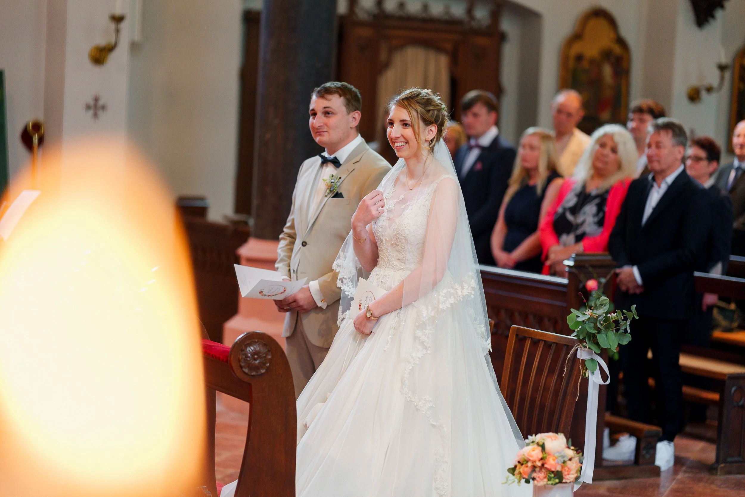 Braut und Bräutigam stehen in einer Kirche während ihrer Hochzeitszeremonie, umgeben von Gästen.