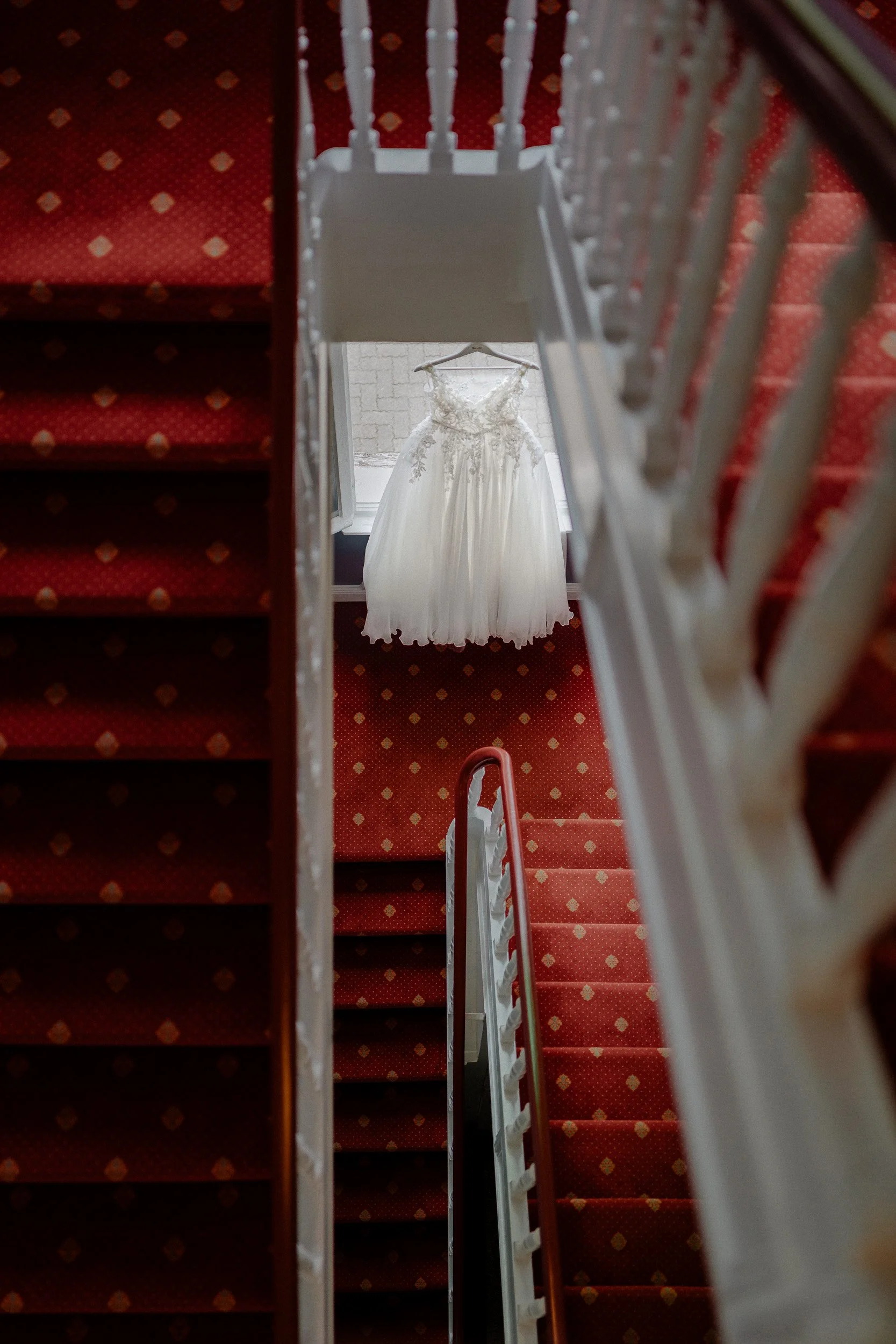 Getting Ready | Gummersbach | Ein Hochzeitskleid hängt an einem Fenster, fotografiert von oben durch die Treppe hinweg, mit roter Teppichboden