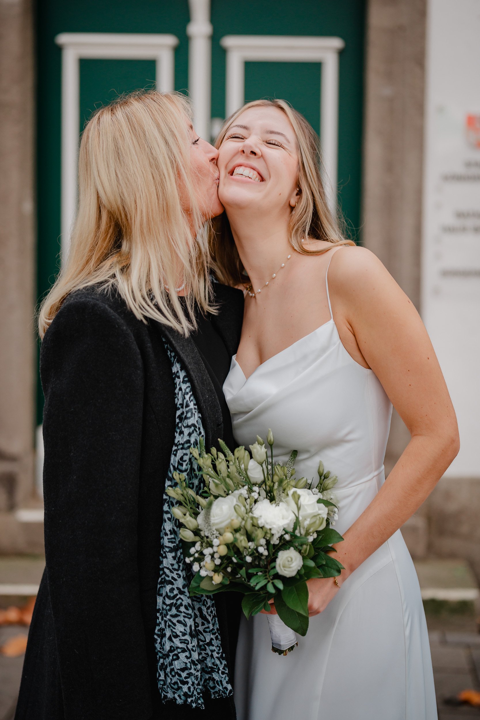 Zwei Frauen, eine in einem weißen Kleid mit Blumenstrauß, küssen sich auf die Wange, Freude bei einer Hochzeit